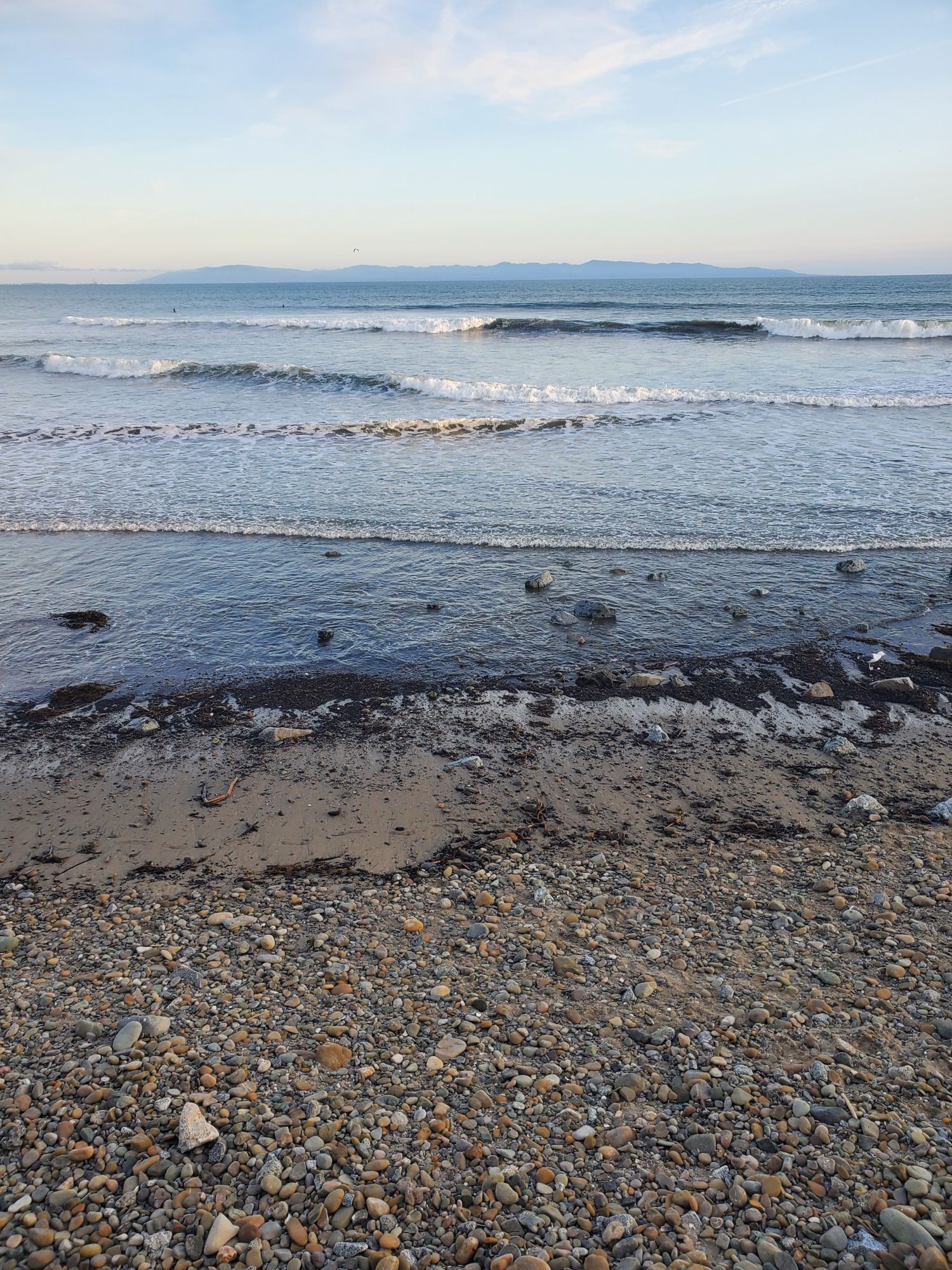 View facing South on a stone crusted slice of Capitola Beach with a crown of ocean broken by 4 layers of low waves all breaking in separate staccato beneath the pale blue sky