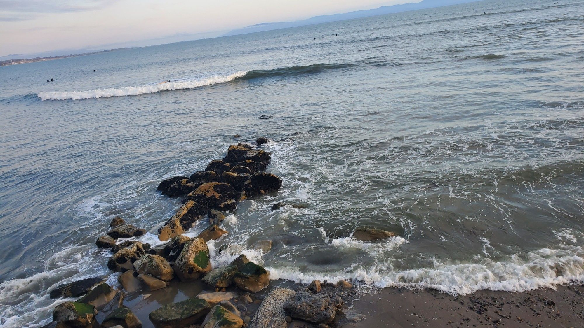 View facing south on Capitola Beach featuring a majority of ocean with a thin wave breaking across the surface. A stone jetty pushes south into the water and it kind of looks like a dude floating on his back with his head facing out to sea and feet towards the sand