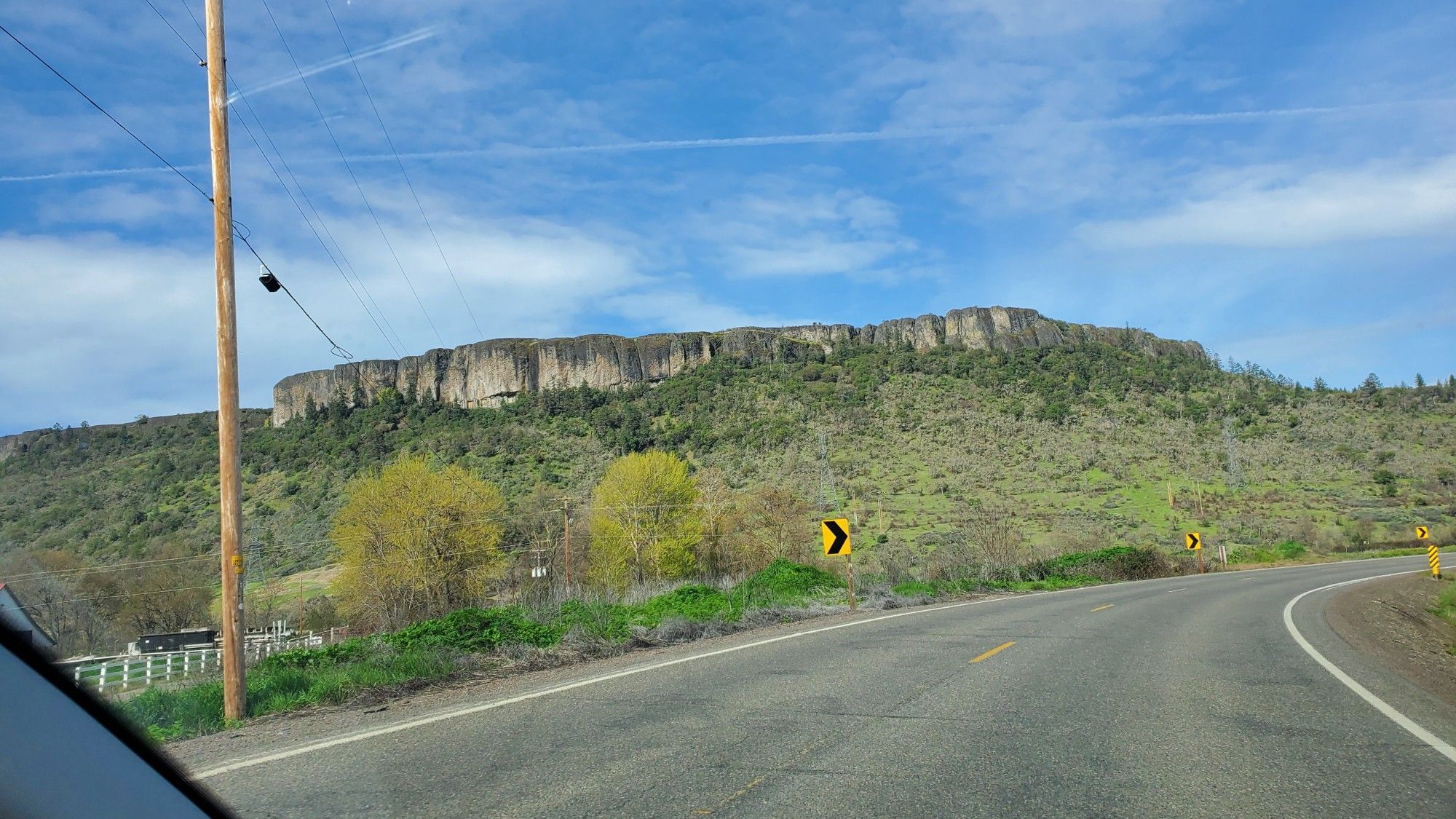 West facing view of Lower Tabletop Rock from the road

Features a wide vibrant blue sky dappled with white clouds over a long plateau of sheer faced stone. The tabletop stands high above rolling hills that are painted with an admixture of evergreen trees closest to the cliff face sloping down to shorter scrub brush and grassland. 

A curve of road is sweeping out from the bottom of frame to the right side.