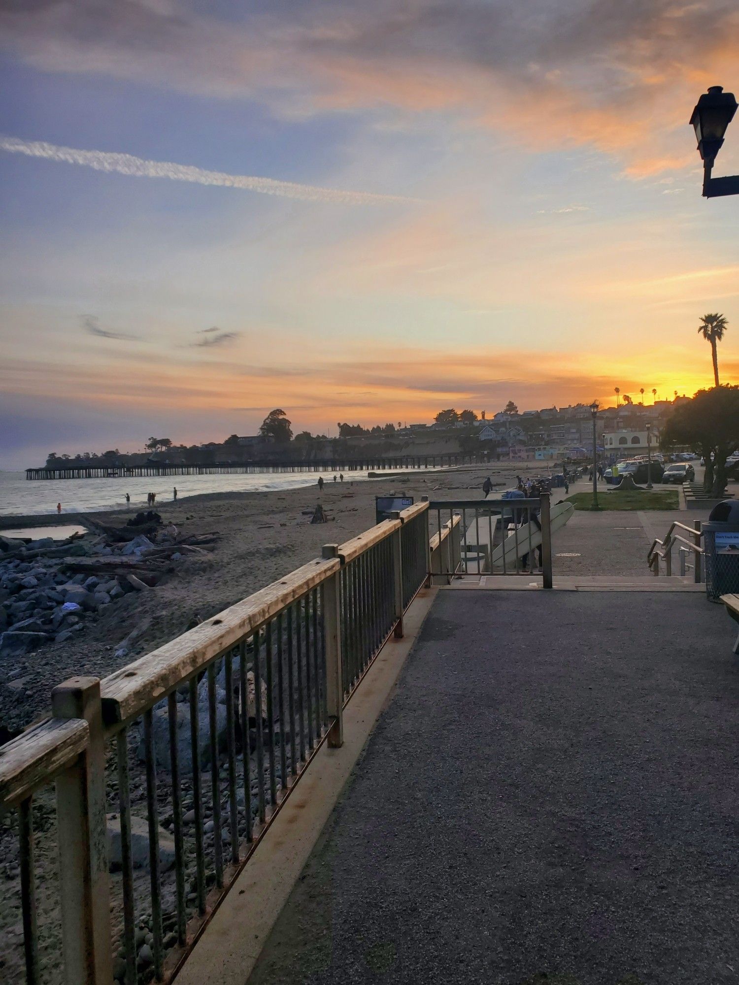 A westward view from Esplanade Park at Capitola Beach. 

Description reads roughly counterclockwise from the upper right corner of frame

Features a wide open sky in a window frame of clouds, with the setting sun over several rows of beachside homes. Outstretched in front of the homes lies a pier running into the water underscored by the ocean and a sweep of sandy beach that is punctuated with stone and driftwood.

A wooden handrail with iron square bars bisects the frame of the photo marching west from the lower left corner out to the center of frame. Finally a paved walkway looking out to the strip of parking and benches dotted with fellow beach goers fills the right hand lower corner of frame.