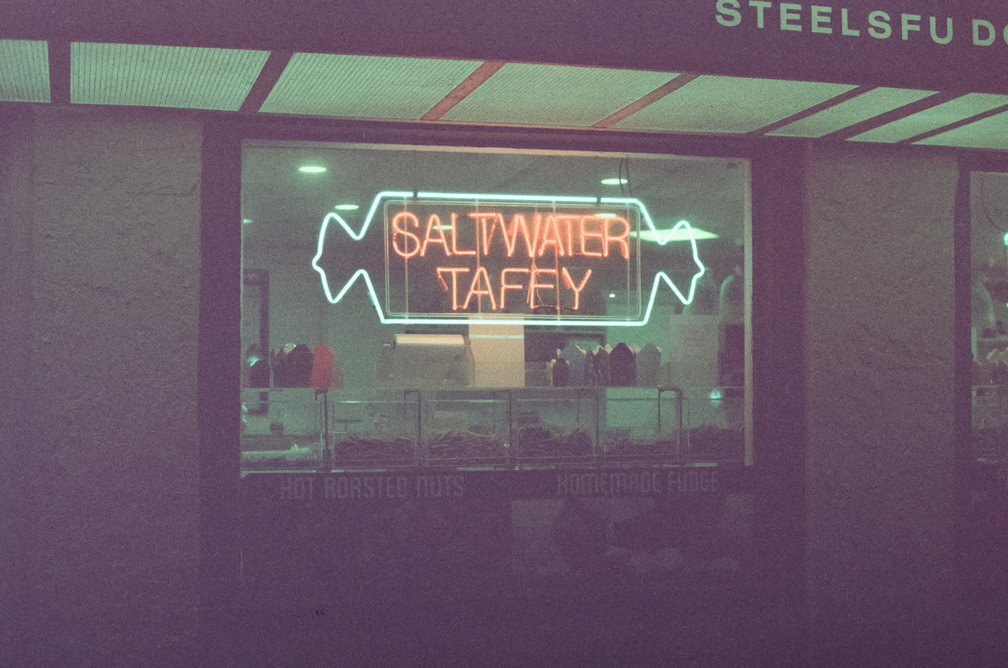 A lit up neon sign that says "saltwater taffy" in the window of Steel's Fudge on a dark Ocean City boardwalk.