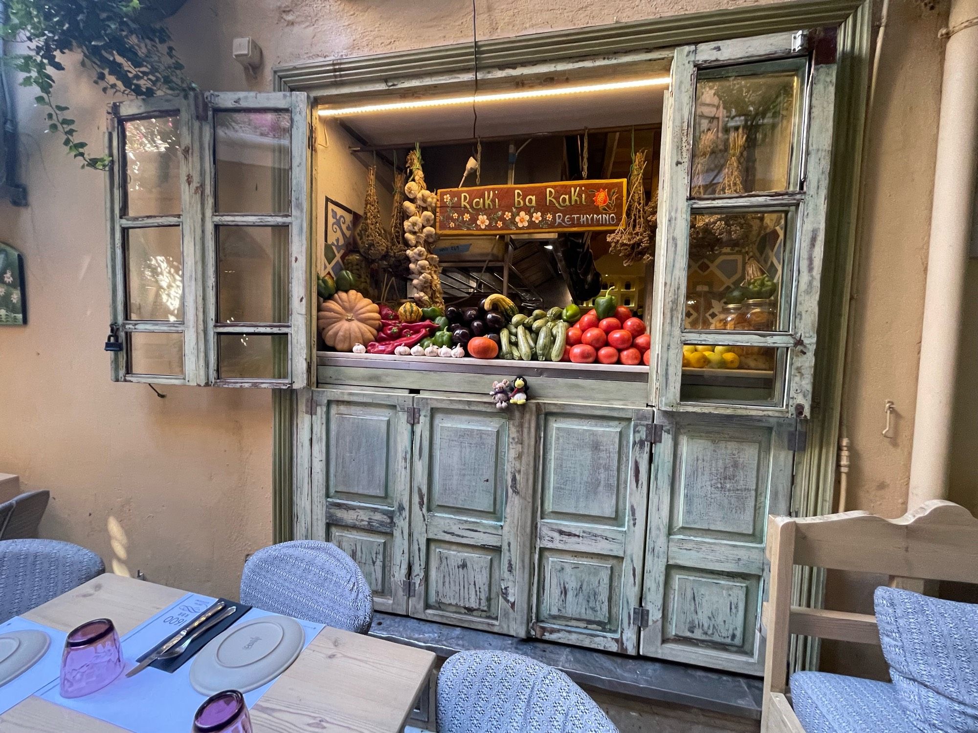 Fenster in der Altstadt von Rethymno auf Kreta. Es gehört zu einem Restaurant, die Fensterflügel sind weit offen und man sieht viel frisches Gemüse, Knoblauch , einen großen Kürbis und im Vordergrund den Plüschpinguin und die Plüschmaus