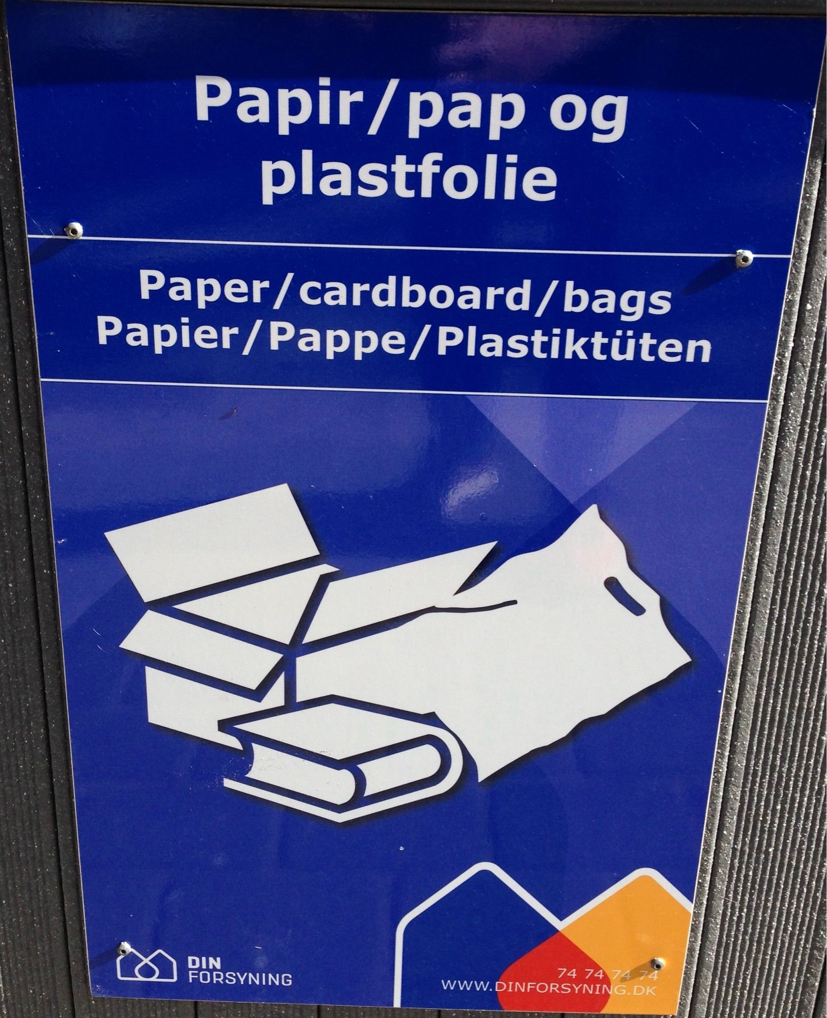 Blaues Schild mit weißer Schrift auf dem dreisprachig (Dänisch/Englisch/Deutsch) auf das gemeinsame Recyceln von Papier und Plastik hingewiesen wird. 
Unter dem Text sieht man in weiß eine grafische Darstellung eines Kartons, eines Buchs und einer Plastiktüte.