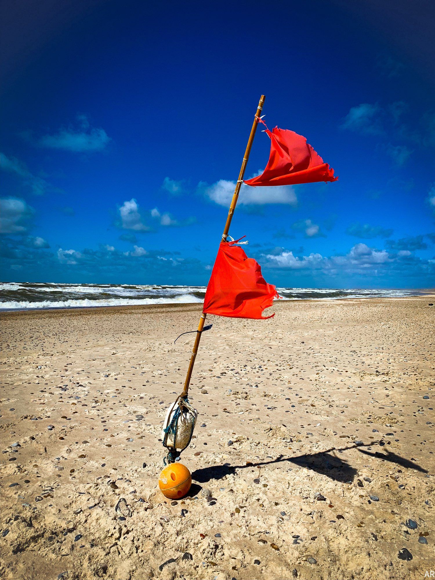 An einem menschenleeren Strand vor blauem Himmel steckt im Bildvordergrund eine Stange mit zwei roten Fahnen, die im Wind wehen. Am Fuß der Stange befindet sich eine fußballgroße, orangefarbene Kugel. Im Hintergrund die aufgewühlte Nordsee.