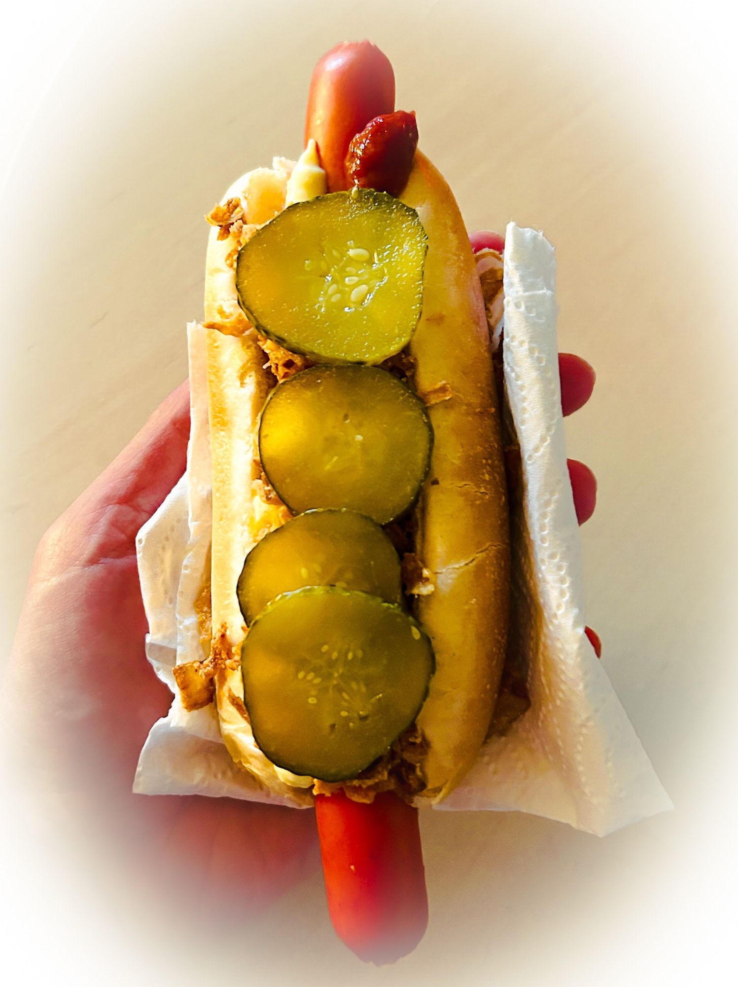 Nahaufnahme eines dänischen Hotdogs, in einer Serviette aufrecht gehalten. Nur echt mit roter Wurst, Röstzwiebeln und Gurkenscheiben!