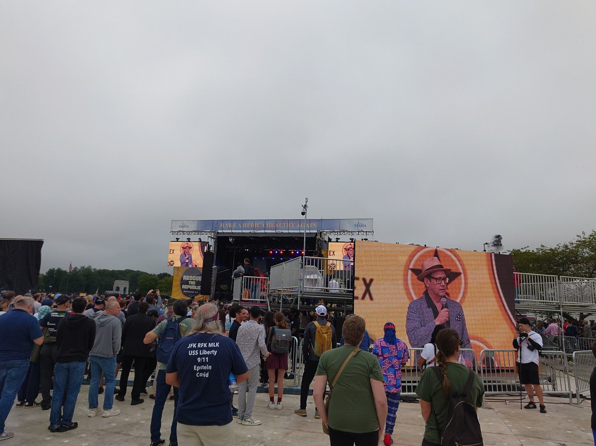 A view of the stage with Rob Schneider in a stupid hat, two screens one near and one far with him on it, in front of a guy wearing a T-shirt

JFK RFK MLL
USS Liberty 
9/11
Epstein
Covid