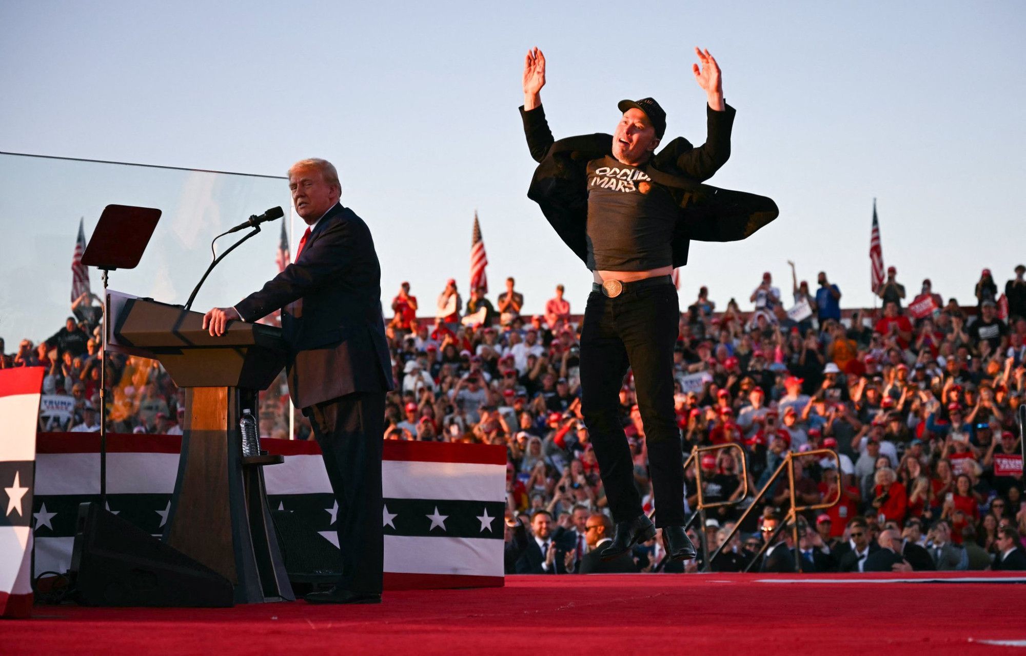 Trump et Musk à une tribune aux USA. Musk fait du trampoline dans sa tête les bras en l'air.