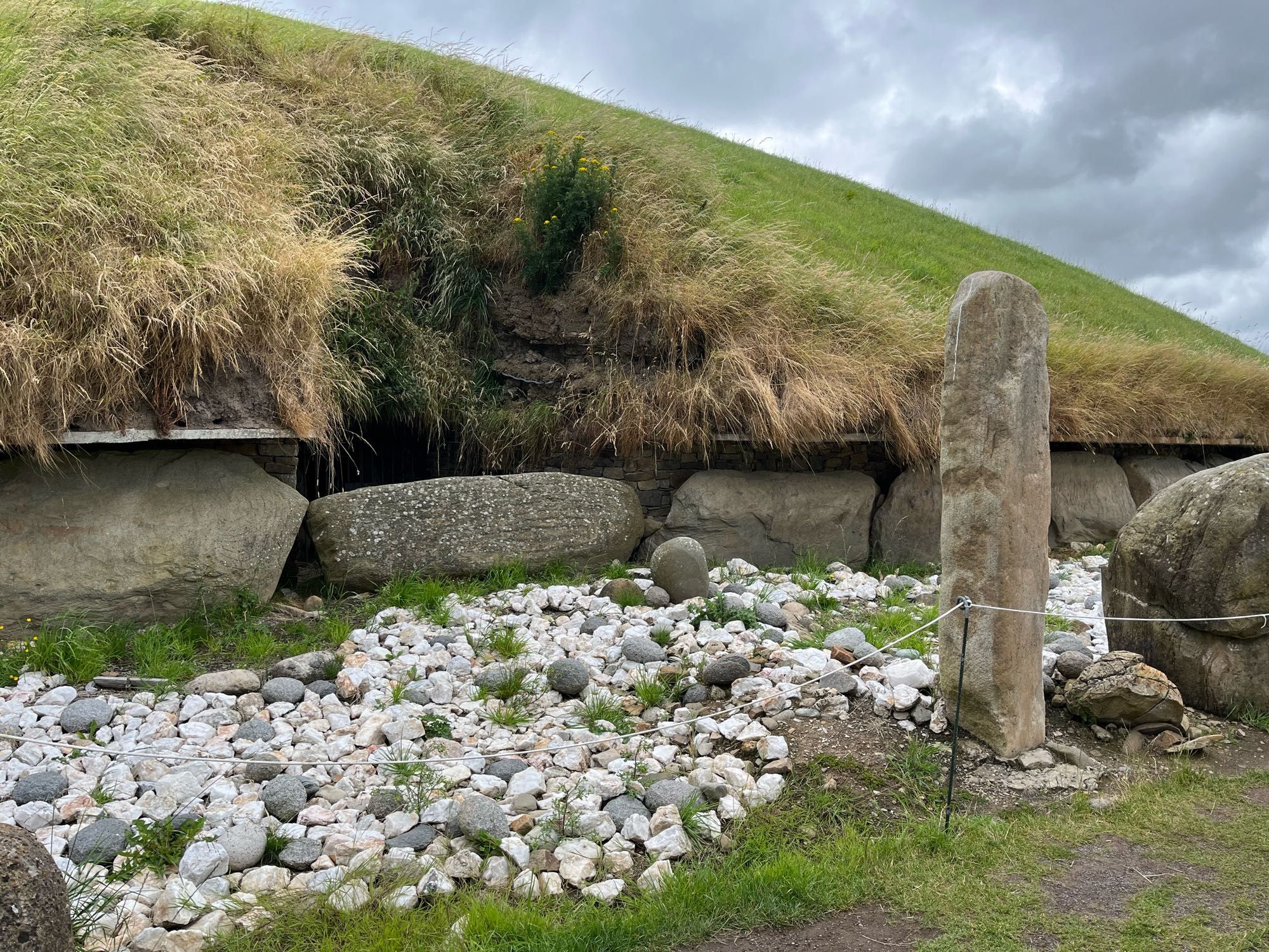 Entrance & standing stone at entrance of West passage of Knowth mound. The mound is covered in grass, and the region between the standing stone & entrance is mostly covered in large rough quartz stones (white) and fewer grey rounded granite stones of similar size