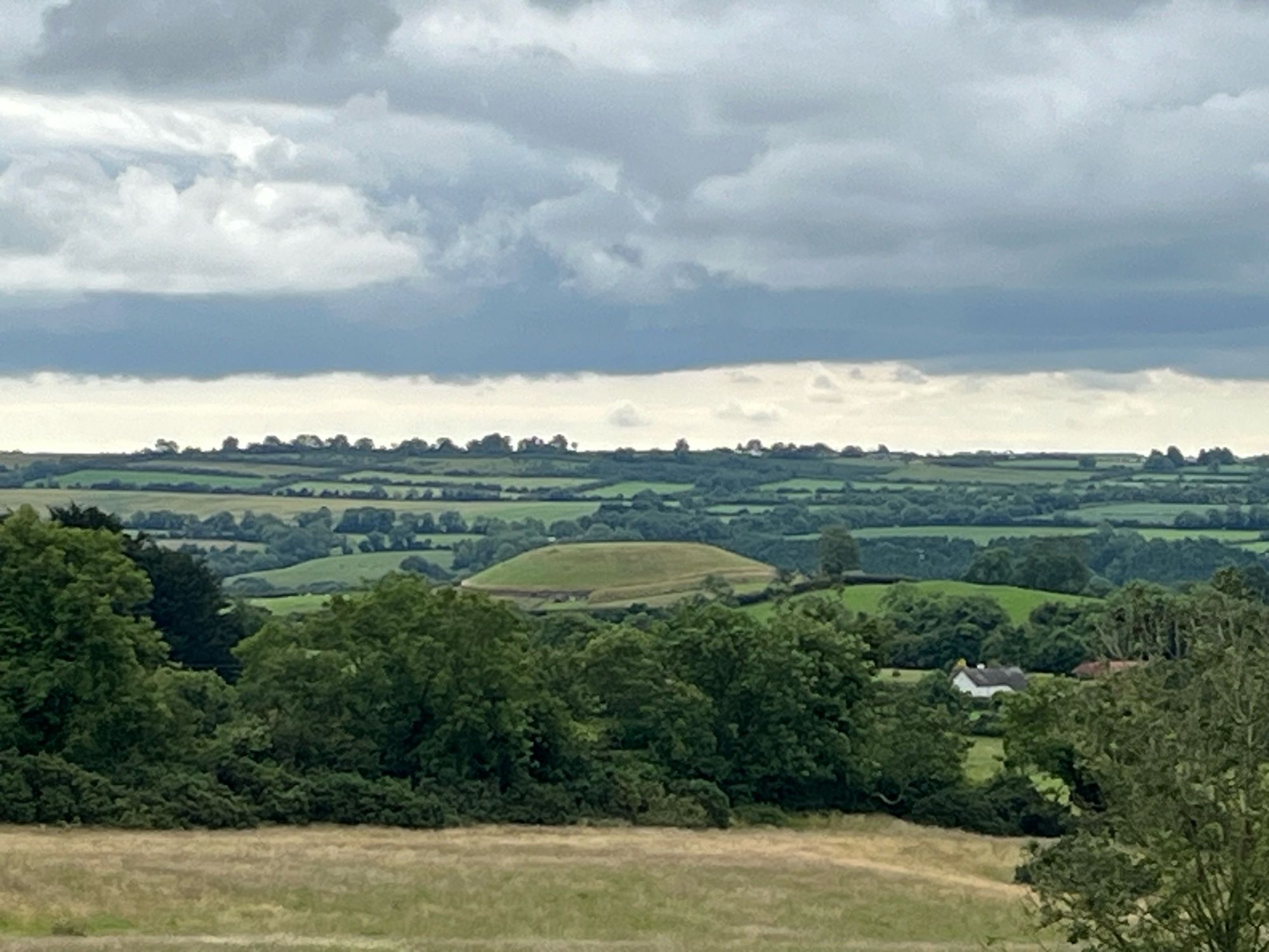 A green landscape of rolling hills with field/hedge system under a grey & threatening sky. A circular, relatively flat-topped mound is visible in the middle distance.