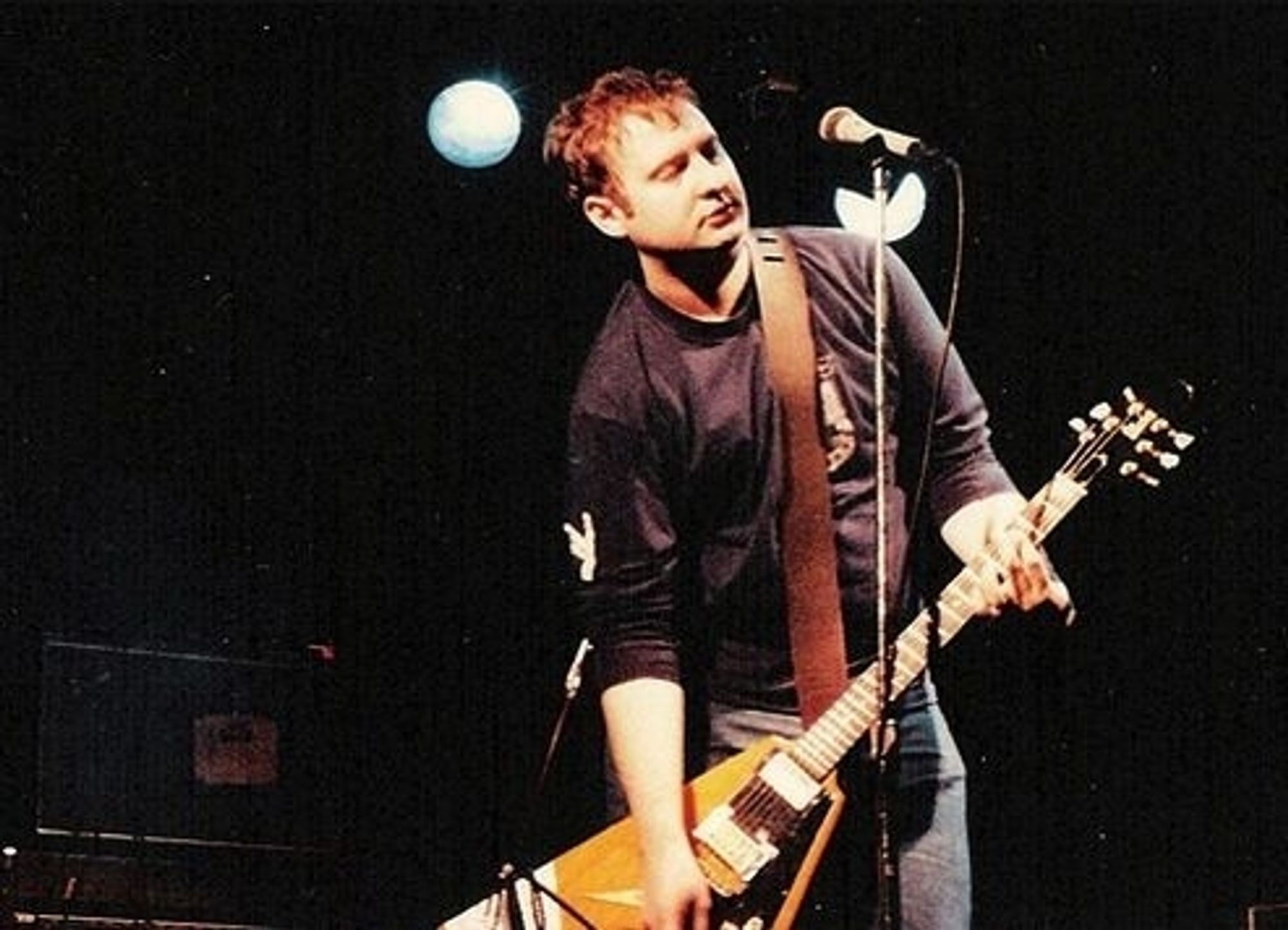 Bob Mould standing at a microphone stand, playing a Gibson Flying V guitar. I'm going to guess sometime in the mid 1980s.