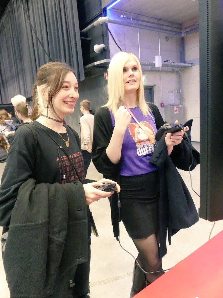 Kora links und Lena rechts an einer PS5 stehend. Kora hat die Haare zum Zopf gebunden, trägt dunkle Kleidung und lacht. Sie hält den Controller in beiden Händen. Lena, die rechte Hand zur Faust geballt, hält den Controller in der linken Hand. Sie trägt ein lila Shirt mit dem Spruch "Bow To Your Queen" und einen schwarzen Minirock. Dahinter sind Menschen und Messestand zu sehen.