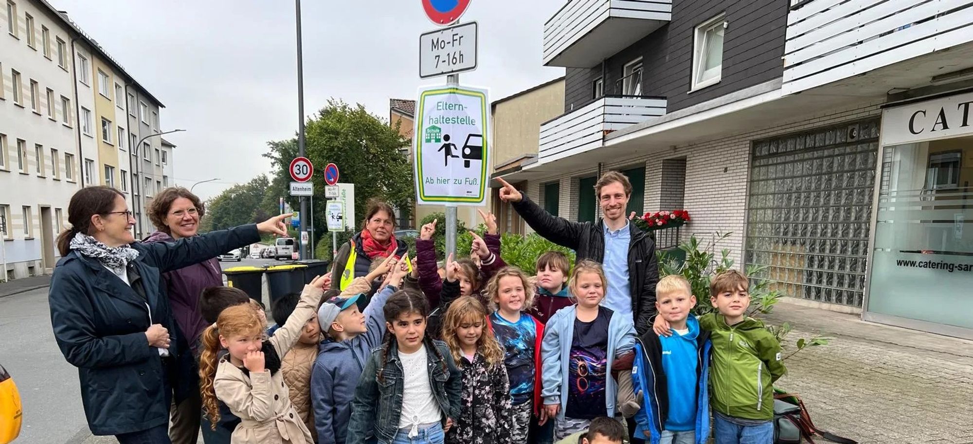 Grundschüler*innen stehen unter einem Schild einer "Elternhaltestelle" und zeigen darauf. Vier Erwachsene tun es ihnen gleich. Foto: Stadt Wuppertal