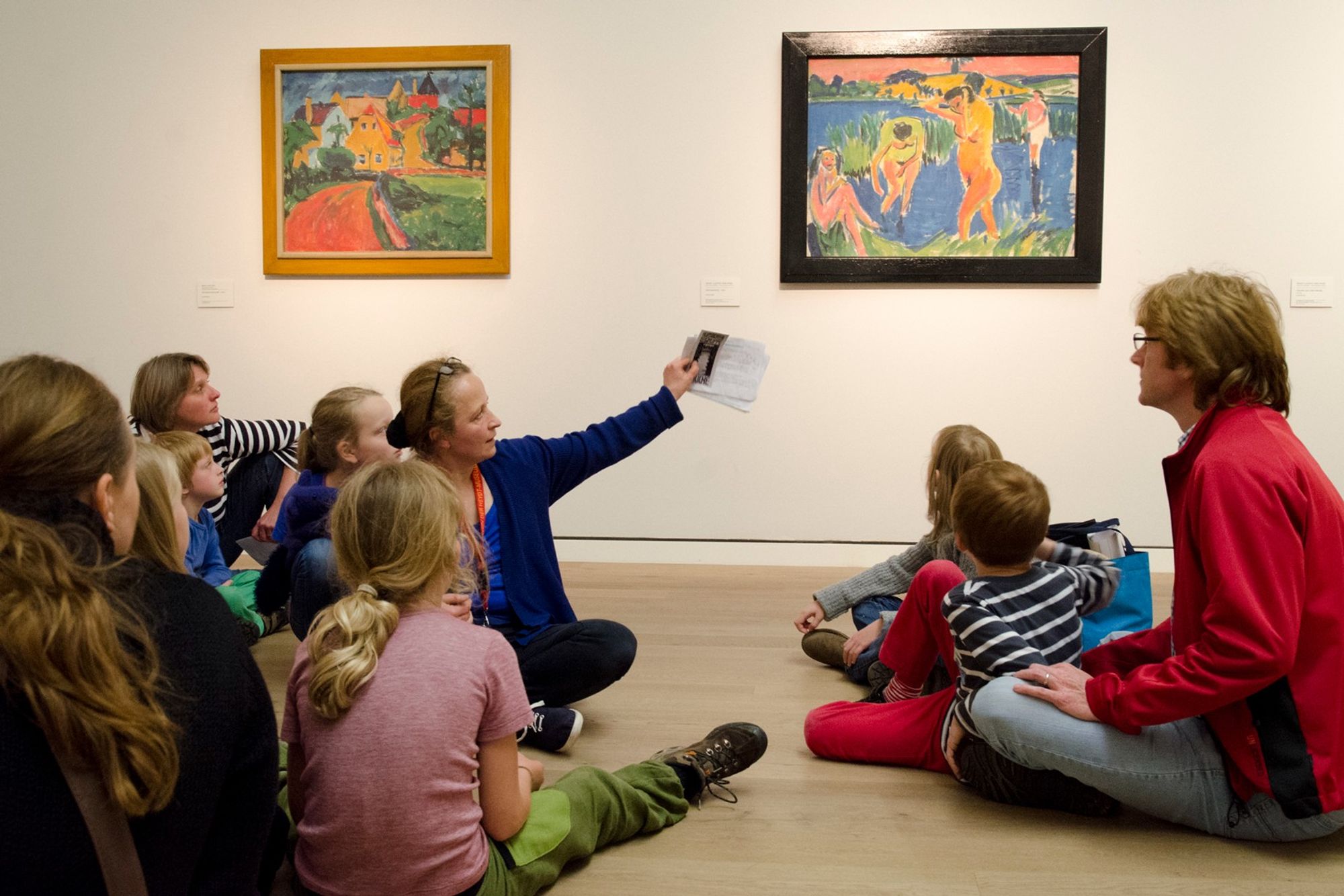 Mehrere Kinder und zwei Erwachsene sitzen vor zwei Gemälden im Von der Heydt-Museum bei einer Familienführung. Quelle: Von der Heydt-Museum