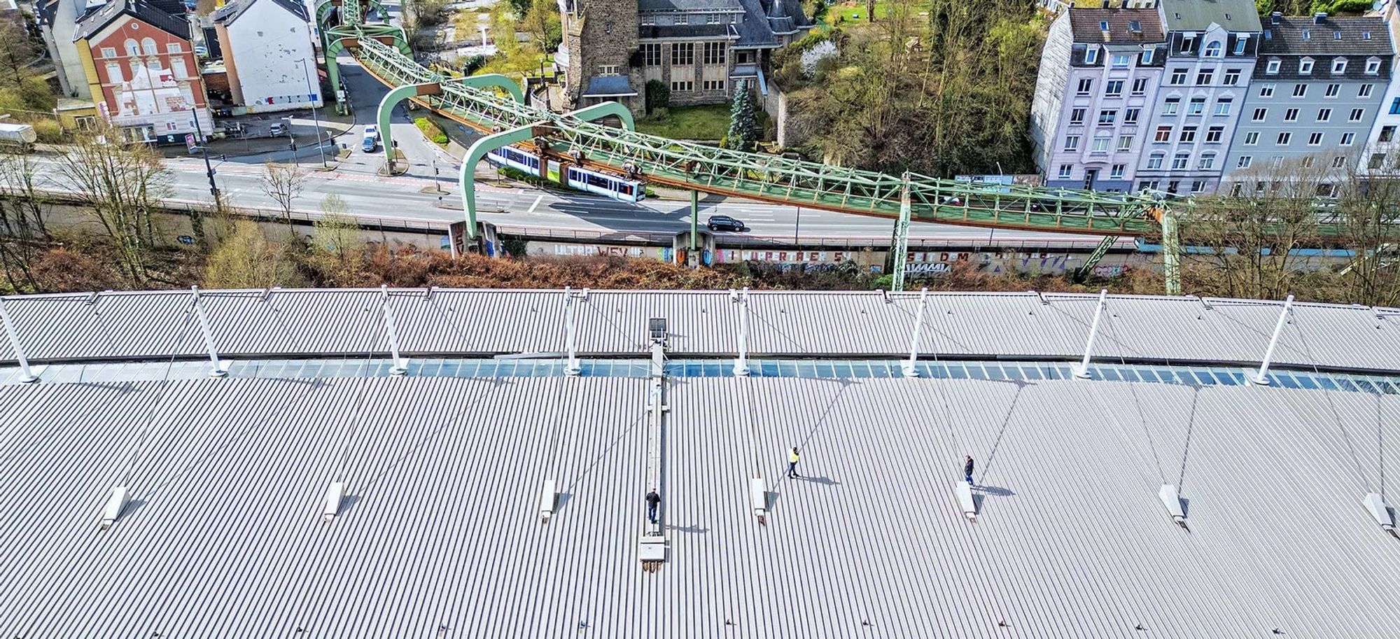 Das noch leere Stadion-Dach von oben, oben im Anschnitt das Gerüst der Schwebebahn. Eine Schwebebahn fährt gerade vorbei, darunter eine Straße mit Bäumen und Häusern. Foto: Buetz, GMW, Stadt Wuppertal