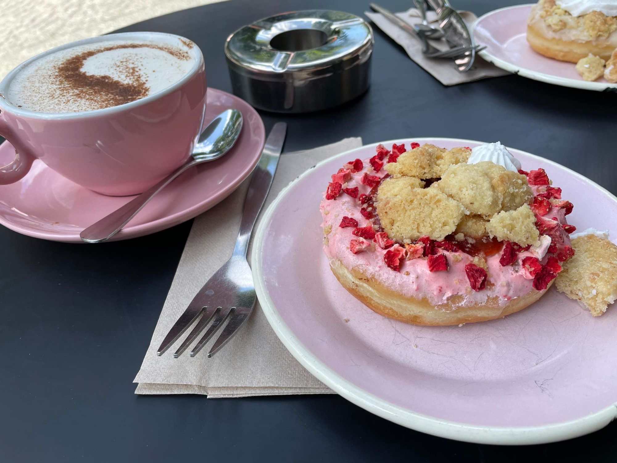 Brammibal's Donuts: rosa Teller mit einem Donut drauf. Rosa Guss, rote Stückchen drauf, außerdem Streusel und ein kleiner Sahneklecks. Links dahinter eine rosa Tasse auf einer rosa Untertasse mit Löffel, in der Tasse Cappucino, oben mit etwas Kakao bestäubt