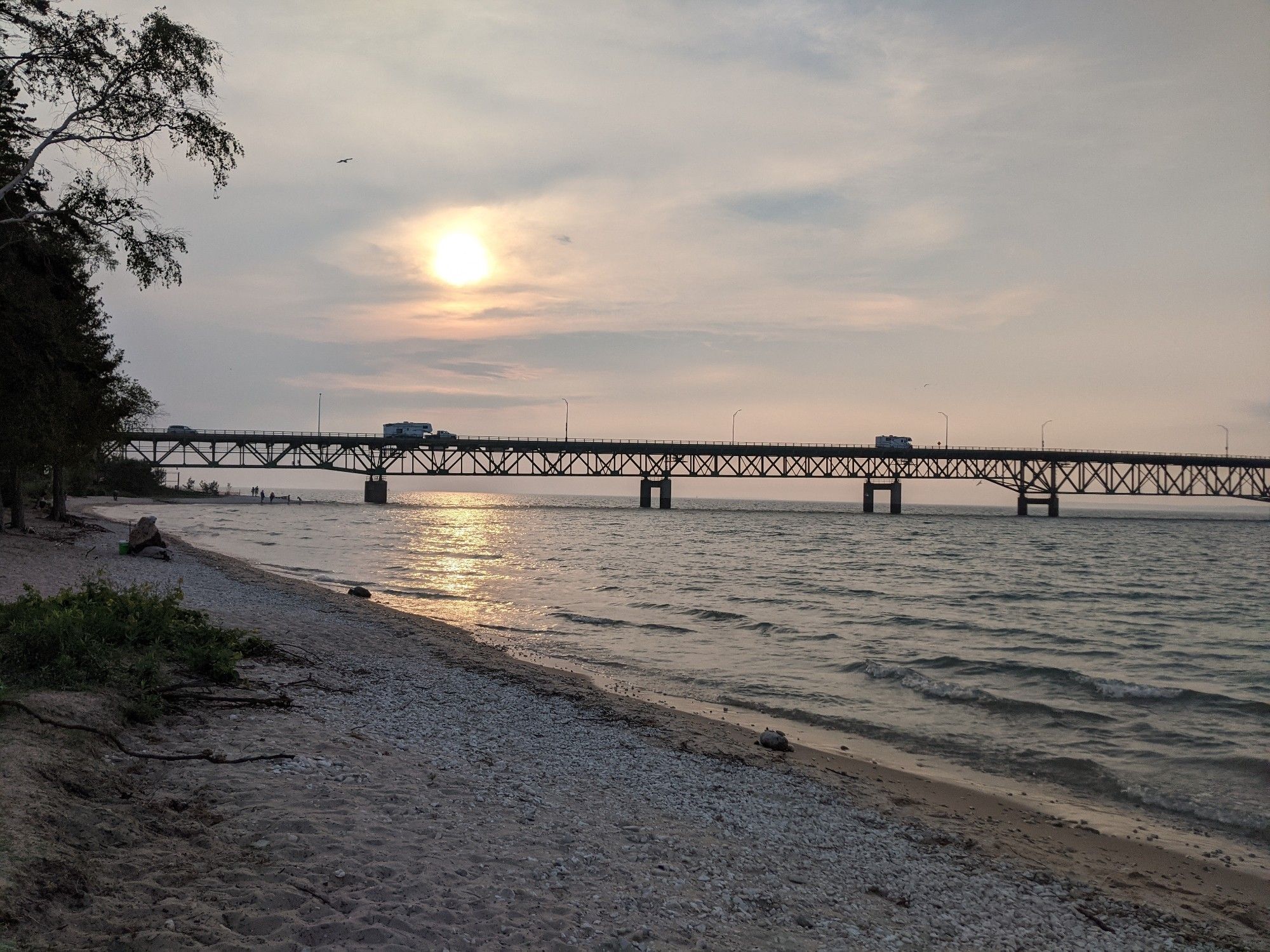 The causeway that leads to the Mackinac Bridge. The sun setting above it. Lake Huron shoreline in the foreground.