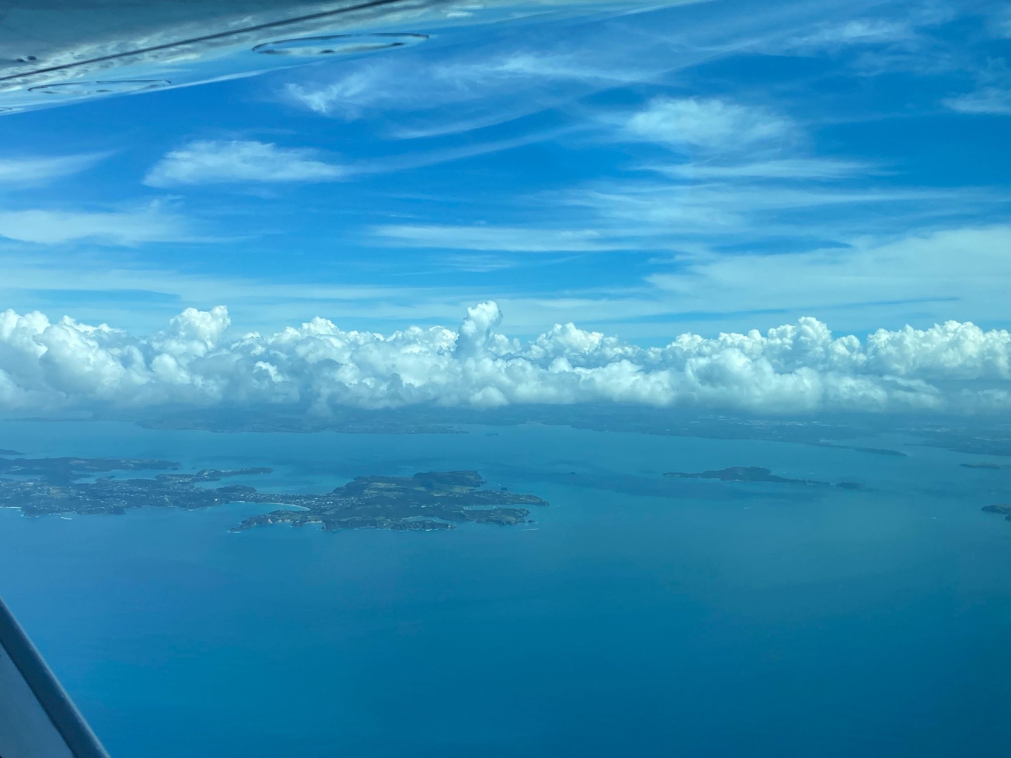 A view across the sea from an aeroplane window, with banks of white clouds hovering above an island. Above the clouds the sky is blue.