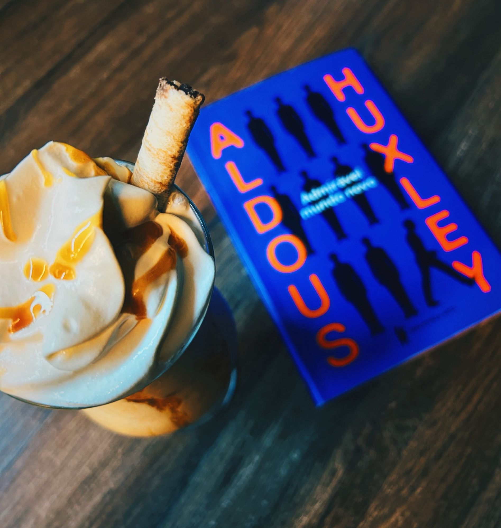 Foto do livro admirável mundo novo em cima de uma mesa de madeira ao lado de um café gelado com chantilly e caramelo em cima numa taça visto de um ângulo de cima meio inclinado. Na capa do livro há várias pessoas igual mas com uma no canto de lado saindo de cena, sua cor ao fundo é roxo.