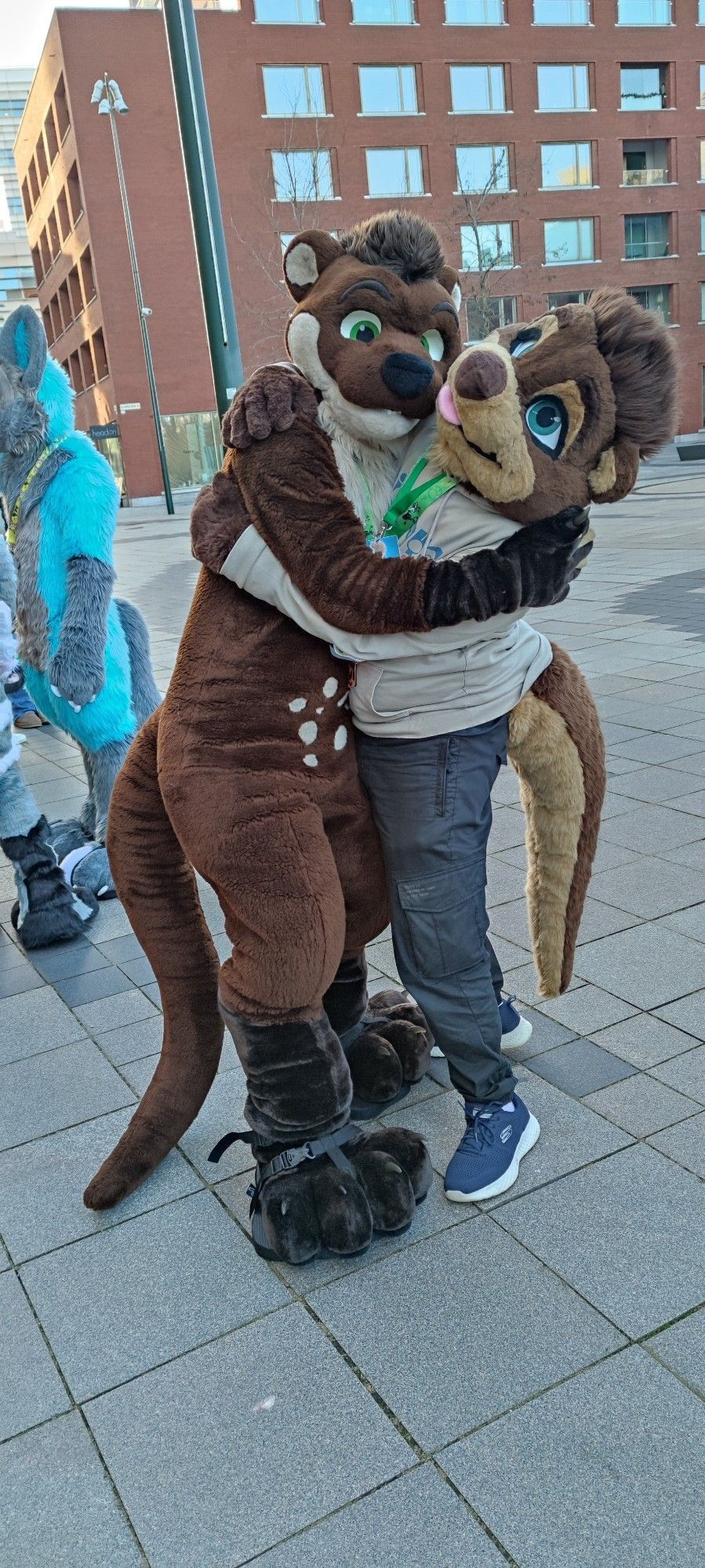 A big, brown and cream colored Otter Fursuiter named Blackymoon hugging another brown and dark brown colored otter Partial-Fursuiter named Jay outside of the Clarion City Live Hotel in Malmö.
Blackymoon has both arms wrapped snugly around Jay as both look towards the viewer into the camera from the side. Blackymoon slightly leans onto Jay as he is taller than him. There are a couple of other Fursuiters standing behind them.