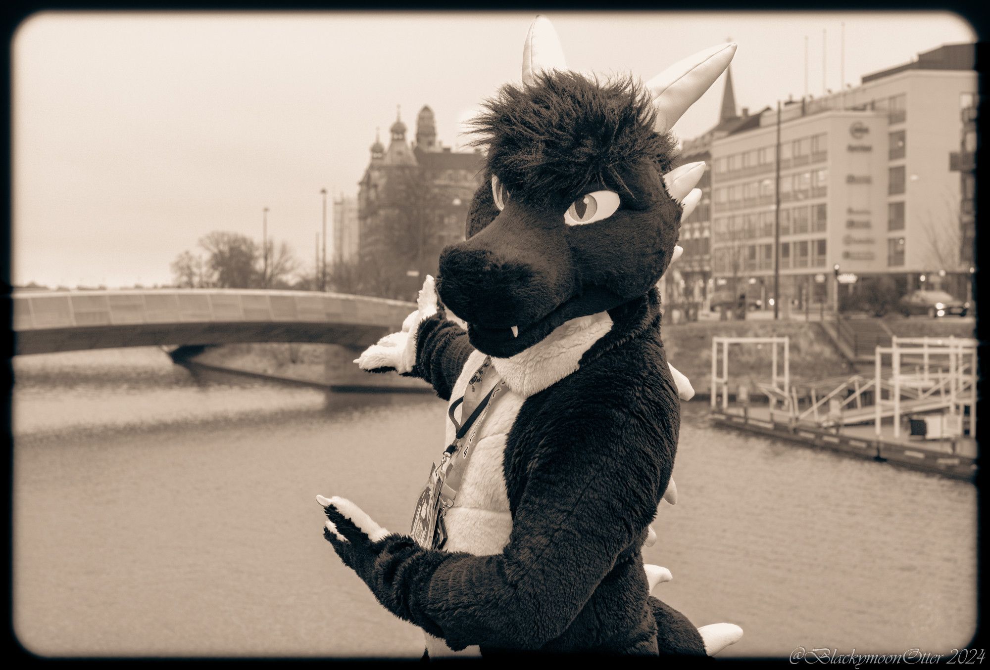 A black and white dragon Fursuiter named Raketh outside the Clarion City Malmö Live Hotel in a portrait shot. He is looking at the camera while pointing behind him towards a river and bridge. The whole photo is dipped in light-brown'ish colors with a darkened frame to make it appear as if it's very old.