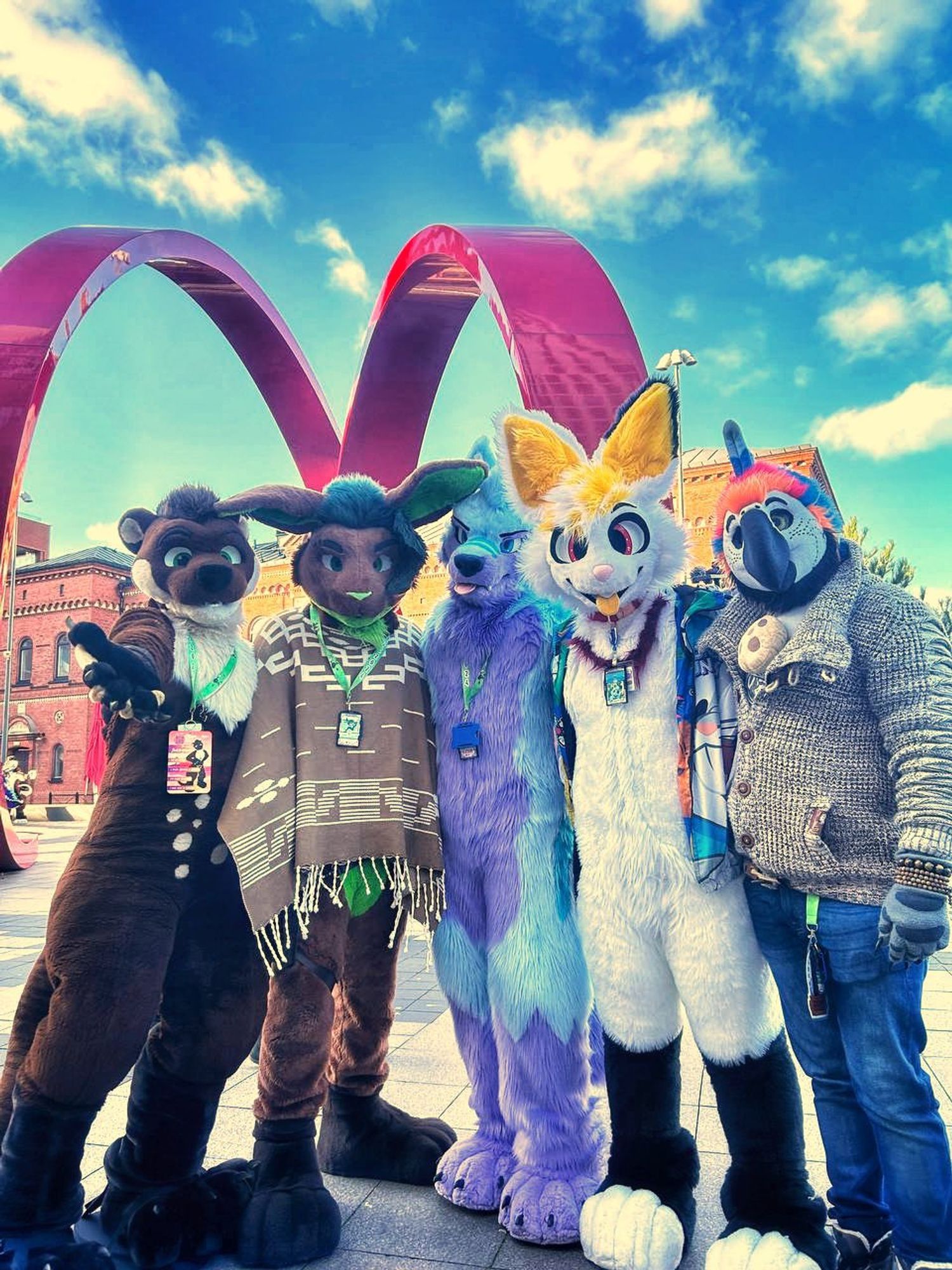 5 Fursuiters (a brown & cream colored Otter named Blackymoon; a brown & green colored bunny named Jinsin; a purple colored wolf named Keenora; a white colored bunny named Fons and a colorful Macaw Partialsuiter named Keks) are standing in front of the giant red curly-statue outside the Malmö Live Hotel in Sweden, looking at the viewer with a nice blue sky in the background.