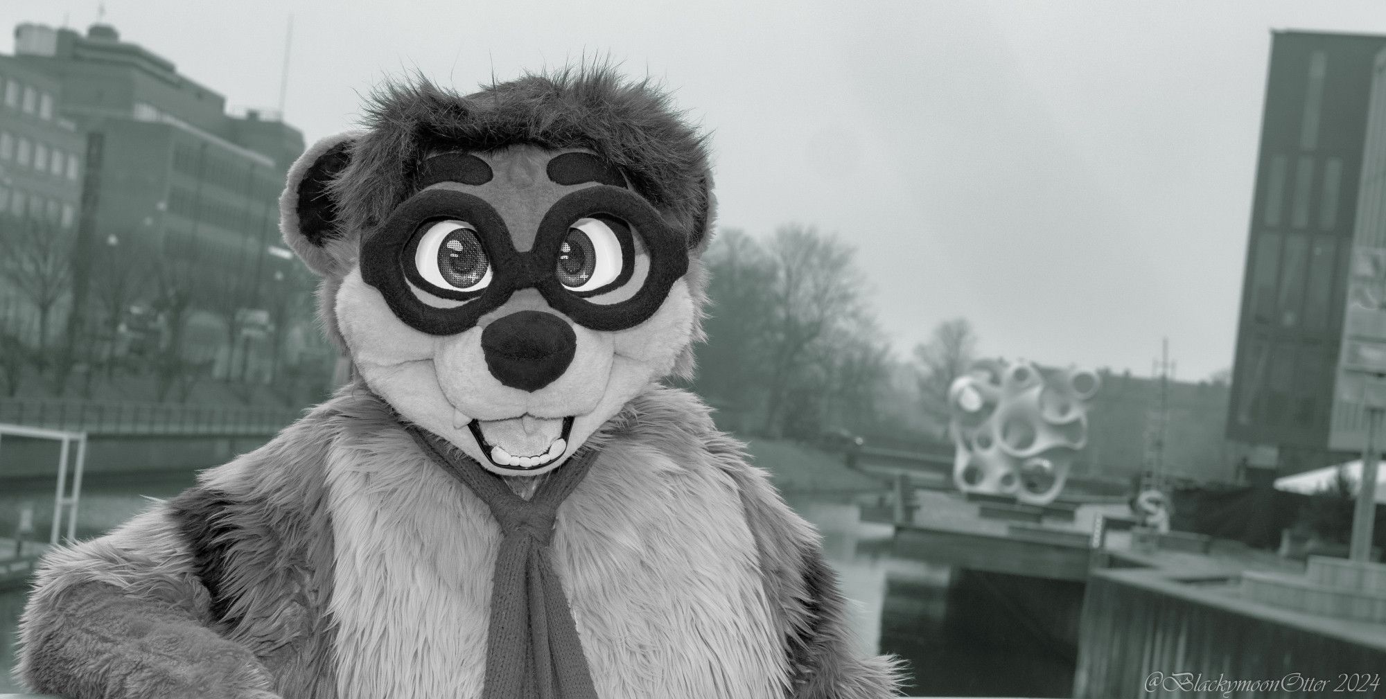 A brown and cream colored Fursuiter named Thabo outside the Clarion City Malmö Live Hotel in a portrait shot, behind him a river and an art statue. Colors were removed from Thabo so he is black & white while the background is a light greenish color.