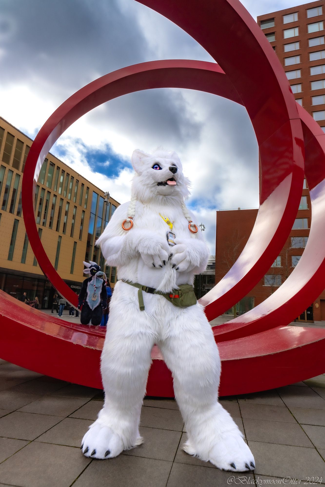 A white canine Fursuiter named Ulven in front of a giant red statue outside the Clarion City Malmö Live Hotel