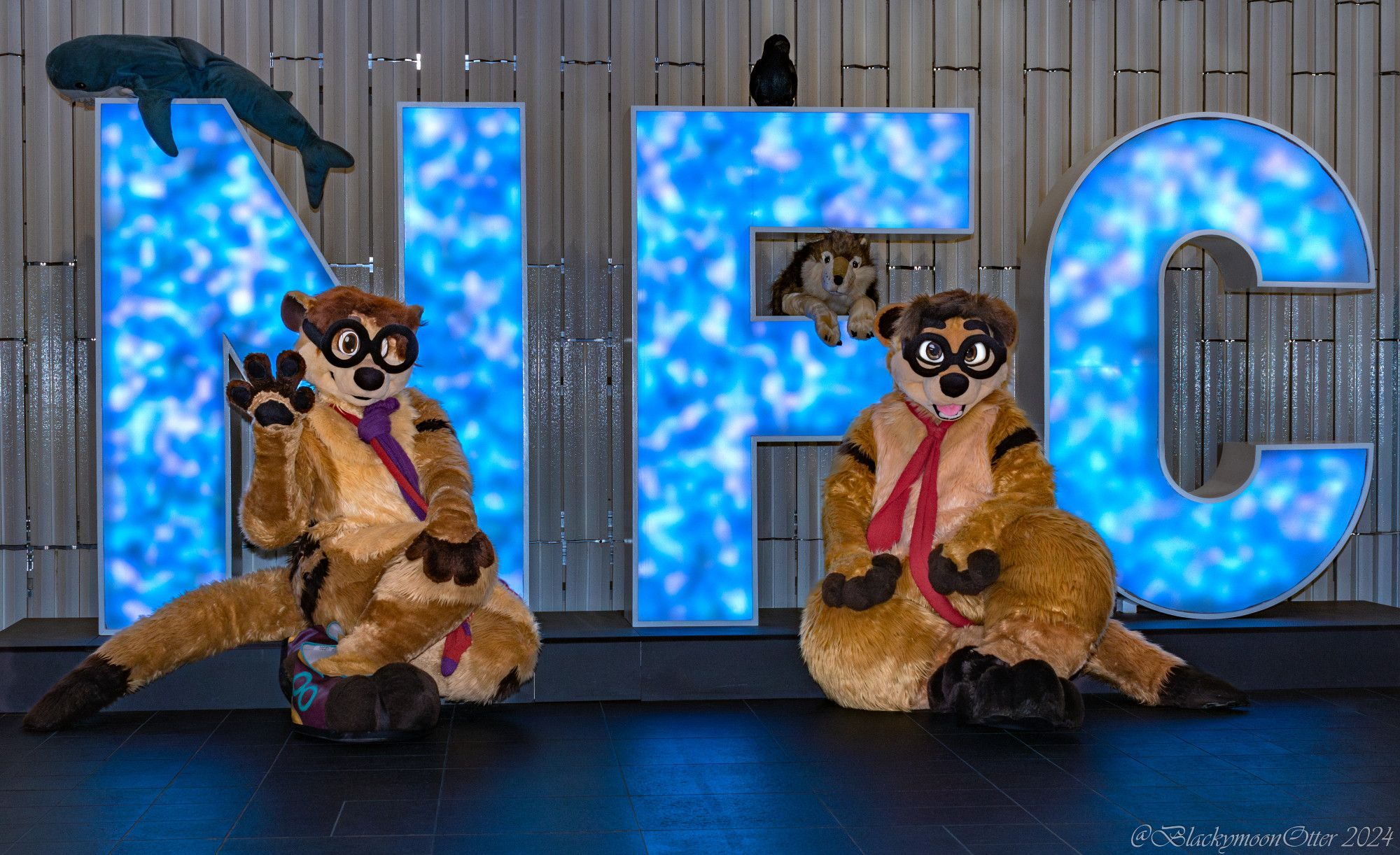 Two brown and cream colored meerkat Fursuiters named Theseus & Thabo kneeling and winking in front of the blue colored NFC Logo inside the Clarion City Malmö Live Hotel.