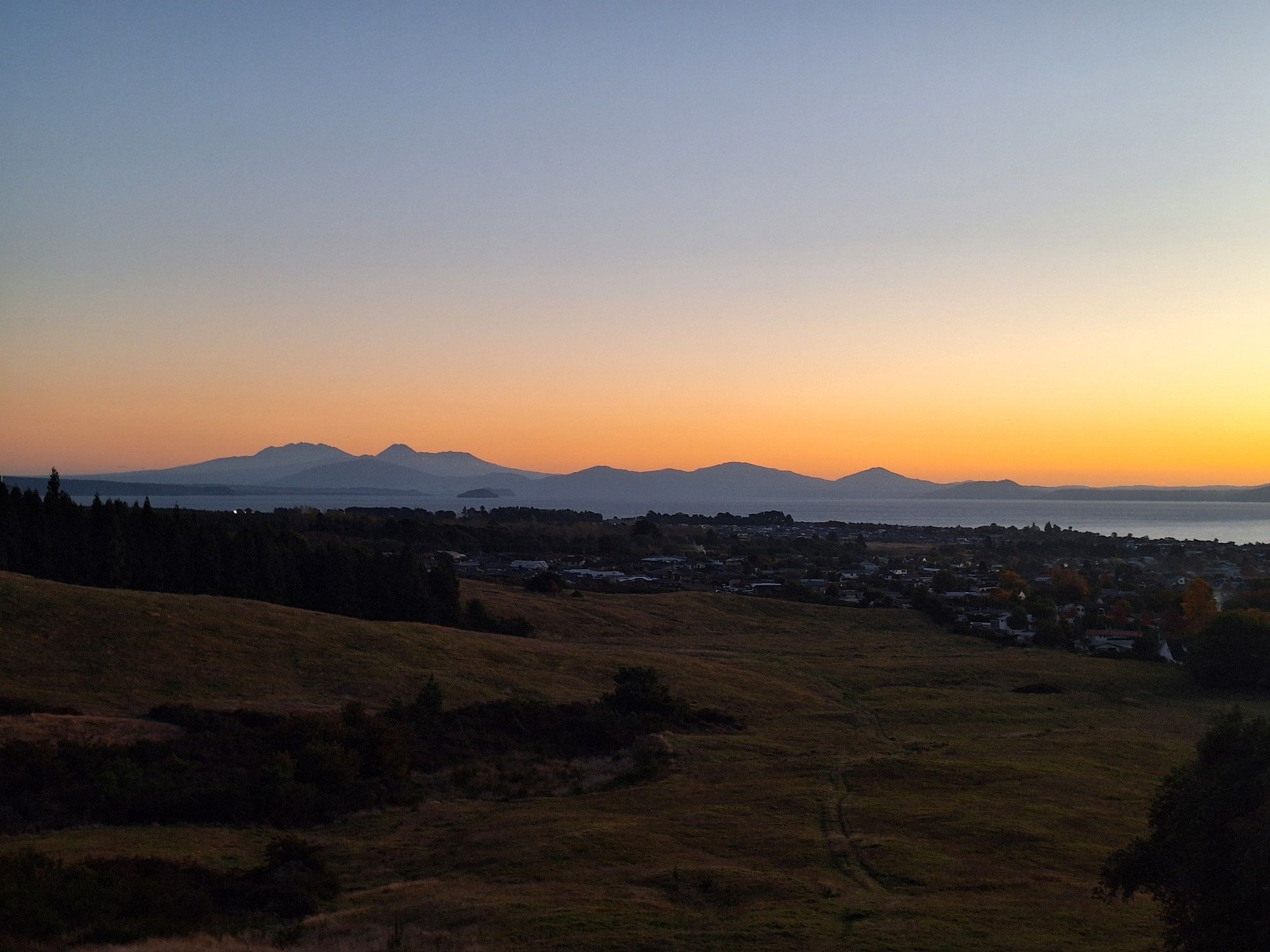 Sunset colours in a clear cloudless sky over a large calm lake. In the distance, a series of volcanoes in silhouette.