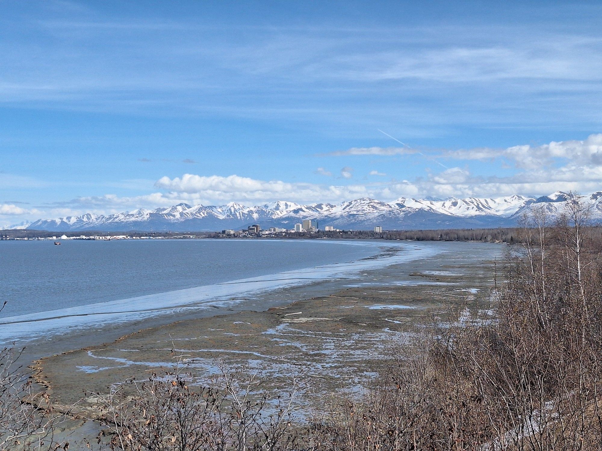 View across a watery scene towards a city skyline, with the backdrop of high and snowy mountains. All under a blue sky with some clouds.