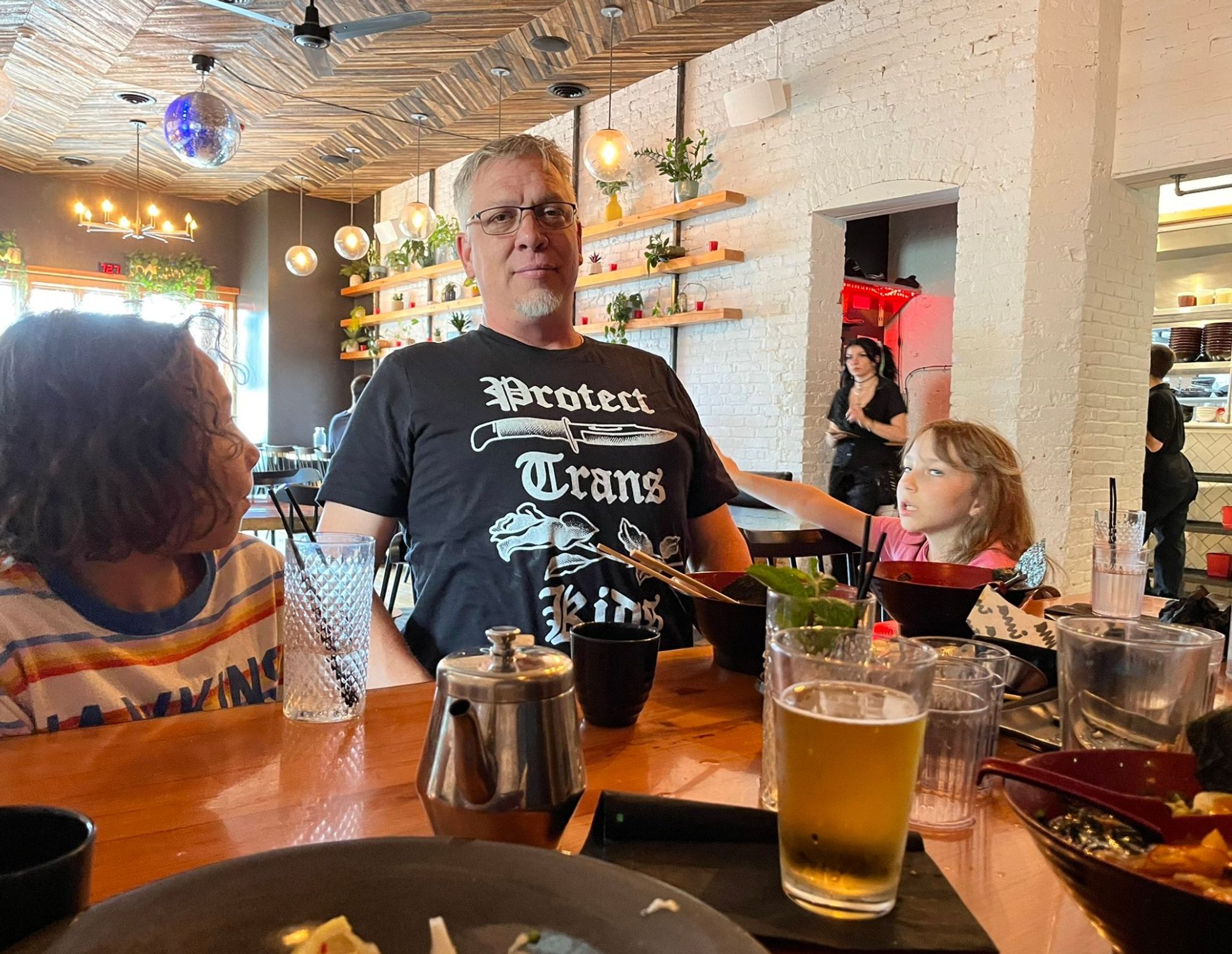 Handsome white guy with glasses and gray goatee sits at a table at a Japanese restaurant with PROTECT TRANS KIDS shirt with big old knife on it. Next to him are a nine-year-old nephew and an eight-year-old daughter who is rolling her eyes until they stick like that. 

Handsome white guy is leaning back and puffing out his chest, the better to show off his awesome shirt. 

Also there's a beer.