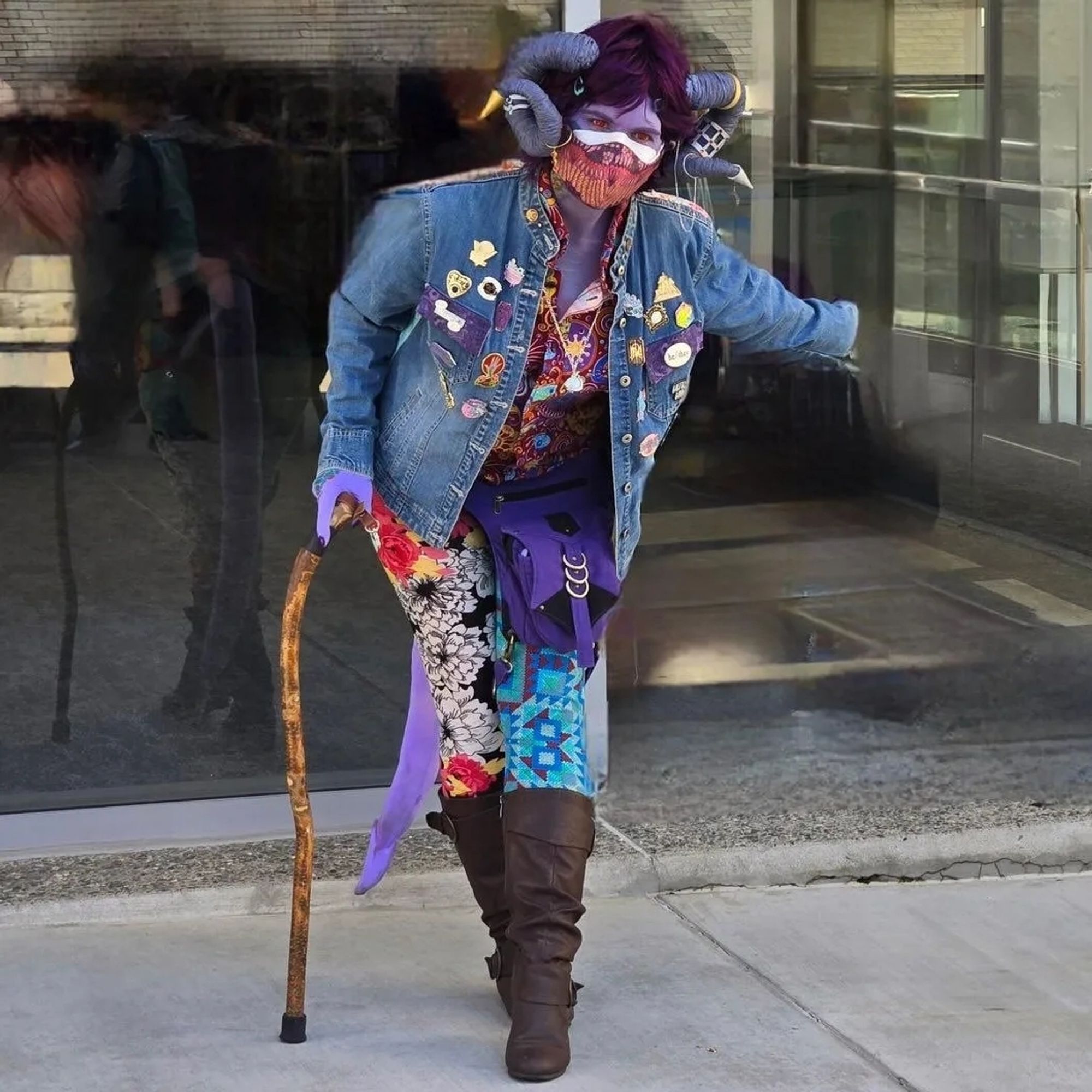 mollymauk cosplayer bows with an arm outstretched. theyre wearing a jean jacket littered in enamel pins and at least four different patterns between face mask, shirt, and leggings.