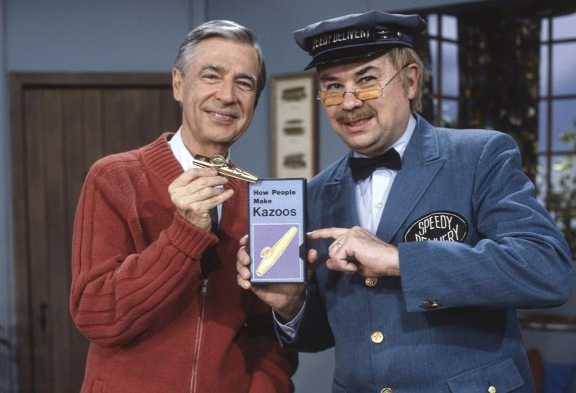 A still image from Mister Rogers Neighborhood in which Mr Rogers, holding a kazoo, stands next to his mail carrier, Mr McFeely, holding a box on which is written, "How People Make Kazoos". They are both smiling and Mr McFeely is pointing at the box.