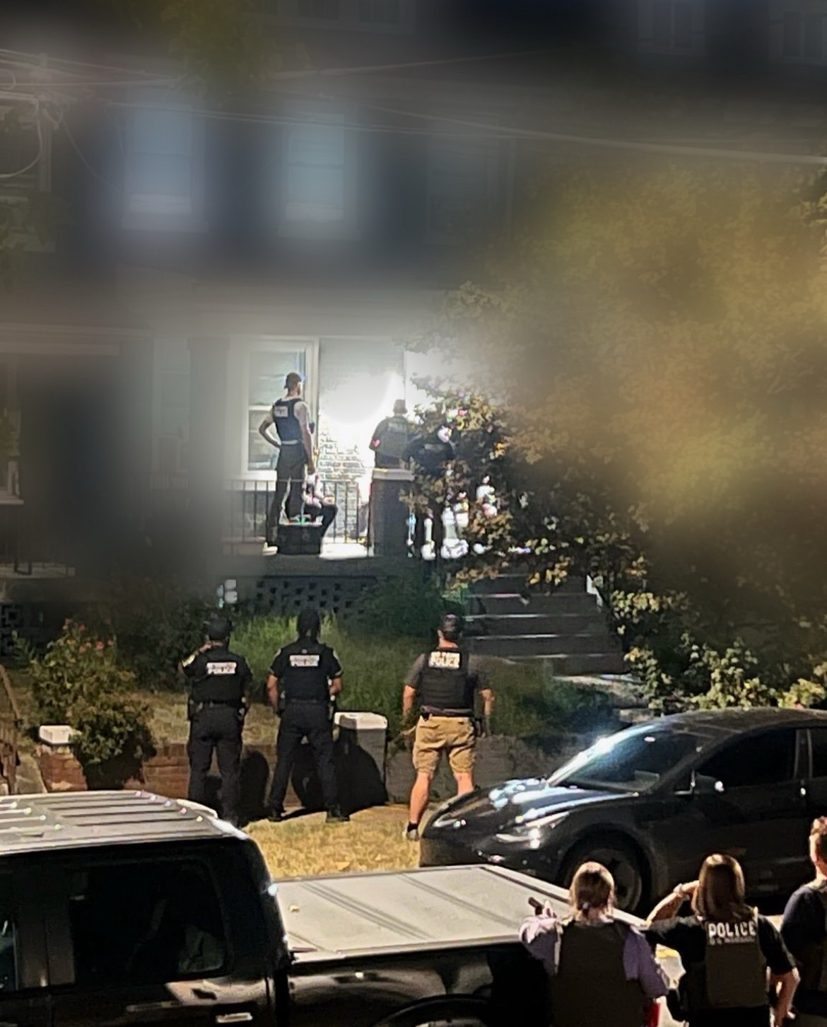 10 cops from MPD, US Marshals, and an unidentified agency likely to be DHS/ICE are standing on a residential street at night. 3 marshals (2 white women, 1 white man) are in the foreground/bottom right of the photo, standing and watching from my side of the street. Across the street, 3 MPD are standing on the sidewalk facing the house/with their backs to me. 4 federal agents are visible on the porch talking to someone who is inside the house, where the front door is open. The background of the other houses is blurred to preserve anonymity and safety.