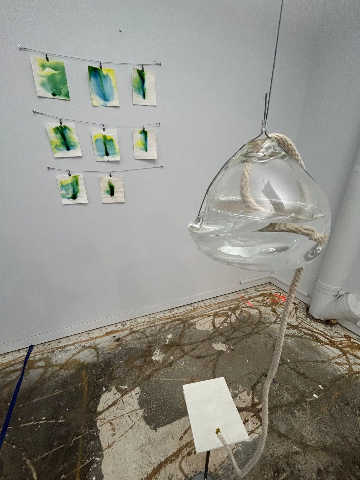 A glass vessel filled with water suspended in the air as a wire. There is a cotton wick rope in the vessel and coming out the hole at the top. The rope moves down to a piece of paper on a stand. In the background there is a series of abstract green images.