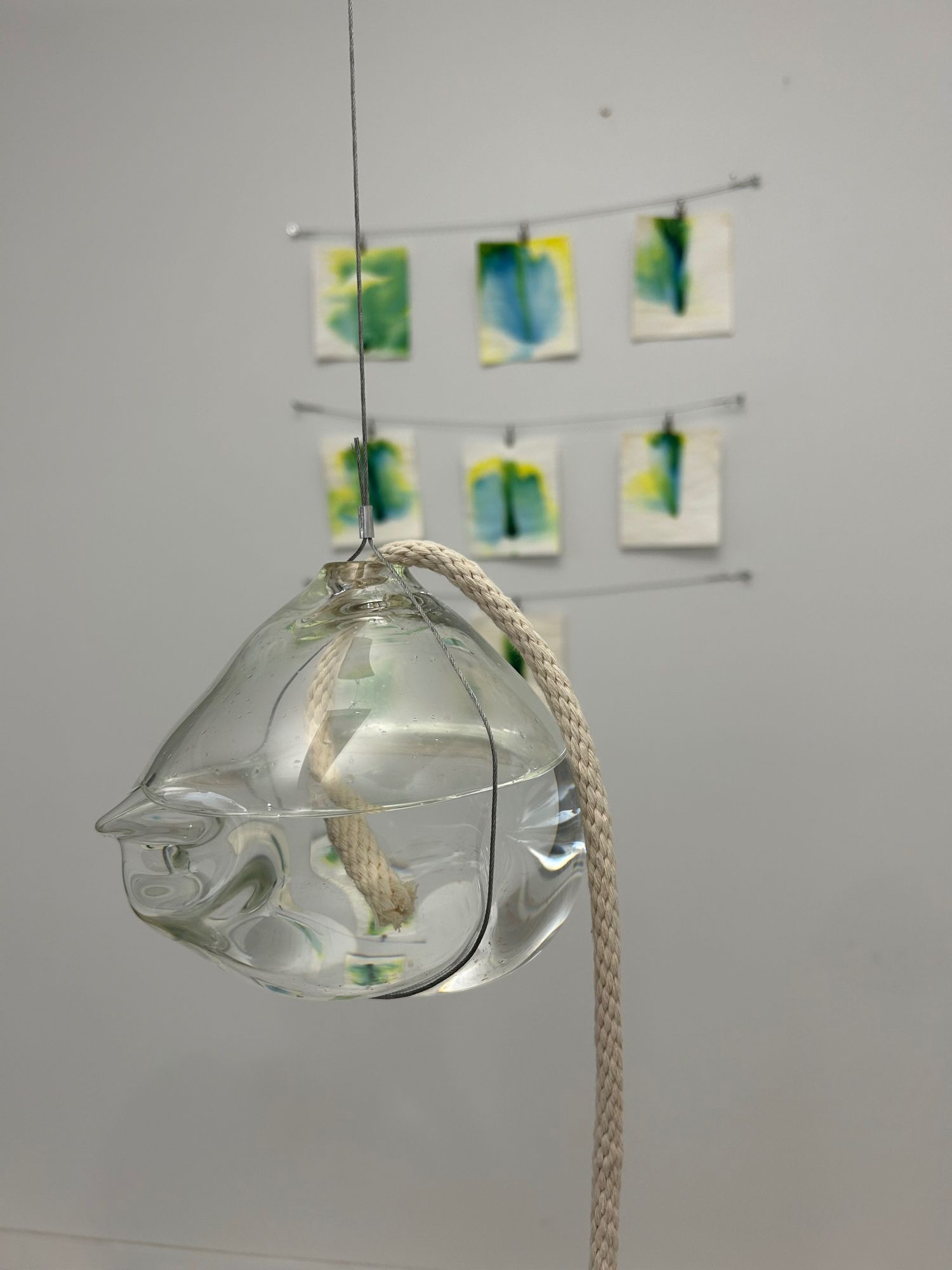 A glass vessel filled with water suspended in the air as a wire. There is a cotton wick rope in the vessel and coming out the hole at the top. In the background there is a series of abstract green images.