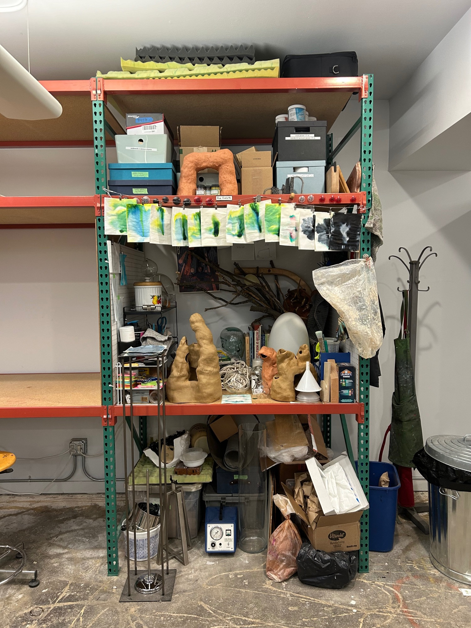 A studio bench that is absolutely stuffed. On the top level are boxes with labels like “teeth” and “tubing.” On the main level are books, sticks, glass and clay sculptures. Below are more boxes and objects including some bags of clay and various air pumps. 