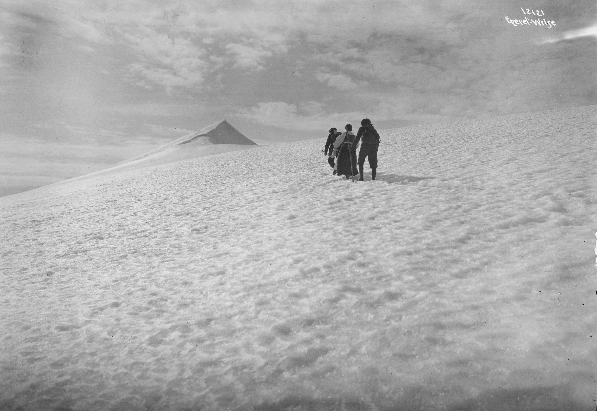 A black and white photograph of three mountaineers from behind, heading across a snow field towards a sharp and dramatic peak on the horizon