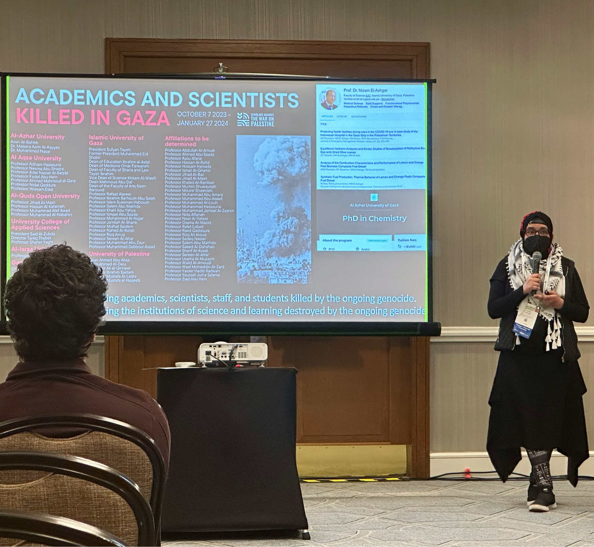 A person wearing a hijab and keffiyeh standing in front of an audience at a conference with a presentation slide behind them. The presentation slide includes a short list of names of academics and scientists who have been killed in the ongoing genocide in Gaza, Palestine (from 10/7/2023-1/27/2024).