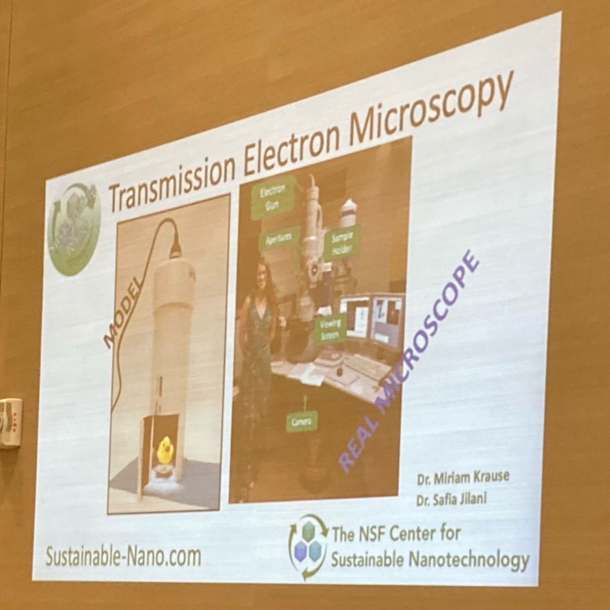 A PowerPoint slide projected on a wall titled: “Transmission Electron Microscopy”

On the left is a model of a TEM that was used in the activity. On the right is an image of a real life TEM with a person standing next to it.