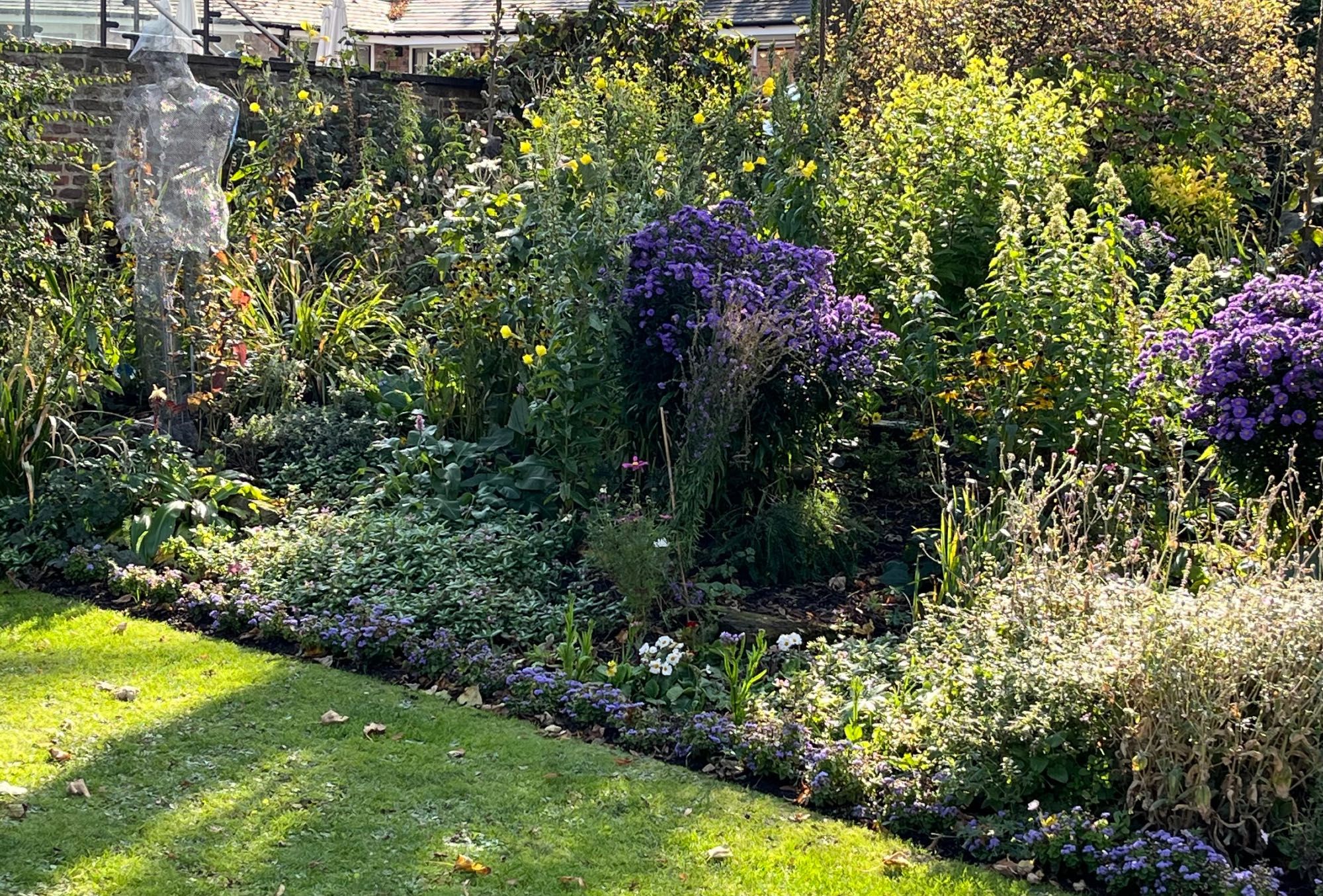 Life size wire work figure of an old fashioned gardener in a flat cap, digging in the corner of a sunny border, with purple asters flowering and a neat lawn in front