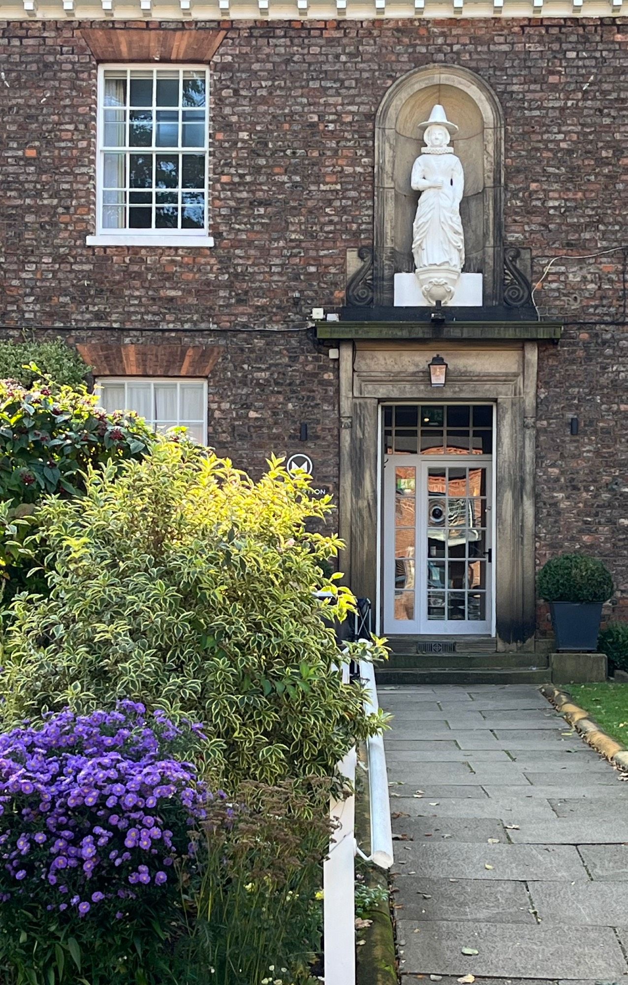 Photo showing rear view of a brickwork hotel, over the door is a large alcove with a 3/4 sized statue of a woman wearing a ruff, a puritan style tall hat and long skirt, in from you can see the end if the garden border and asters flowering by the path