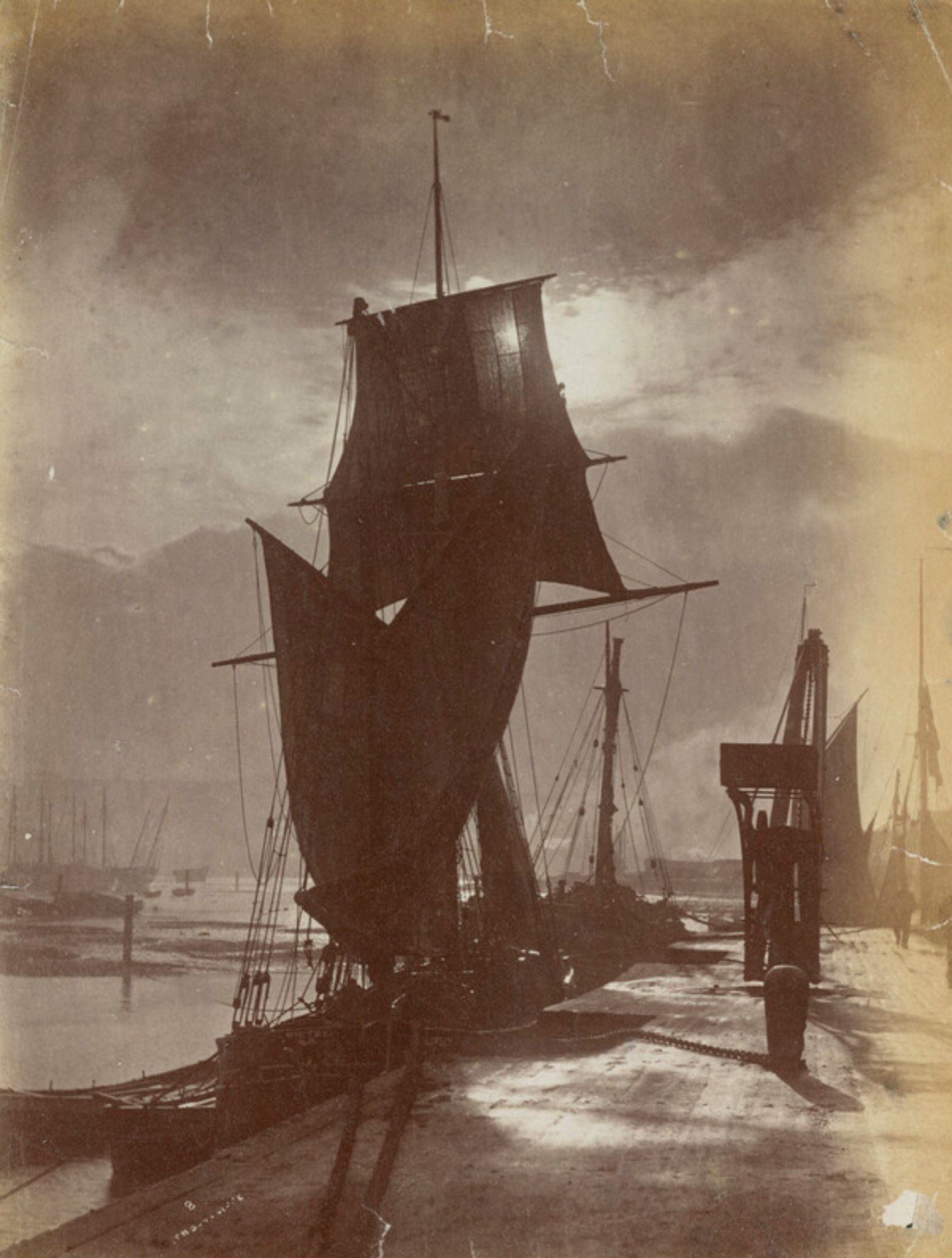 View of a nineteenth fishing vessel from the age of sail, berthed at the quay, a sepia toned print almost expressionist in composition, with sun shining through cloud and sails. Suggestive of a still from Murnau's Nosferatu.