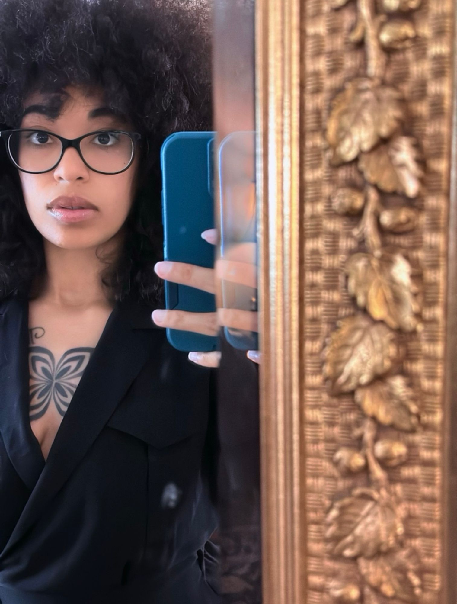 I’m standing in front of a mirror - I’m wearing a black dress that crosses low giving me a long neckline expose my chest and part of my boob, black glasses, and my phone (teal) and some of my fingers are visible but distorted in a sort of “doubled” way (because of the mirror), I have on lip gloss 

chest tattoo visible - I love it 

I look like a dear in headlights 😂 there is a gold mirror edge that’s taking of the right half/ish of the picture