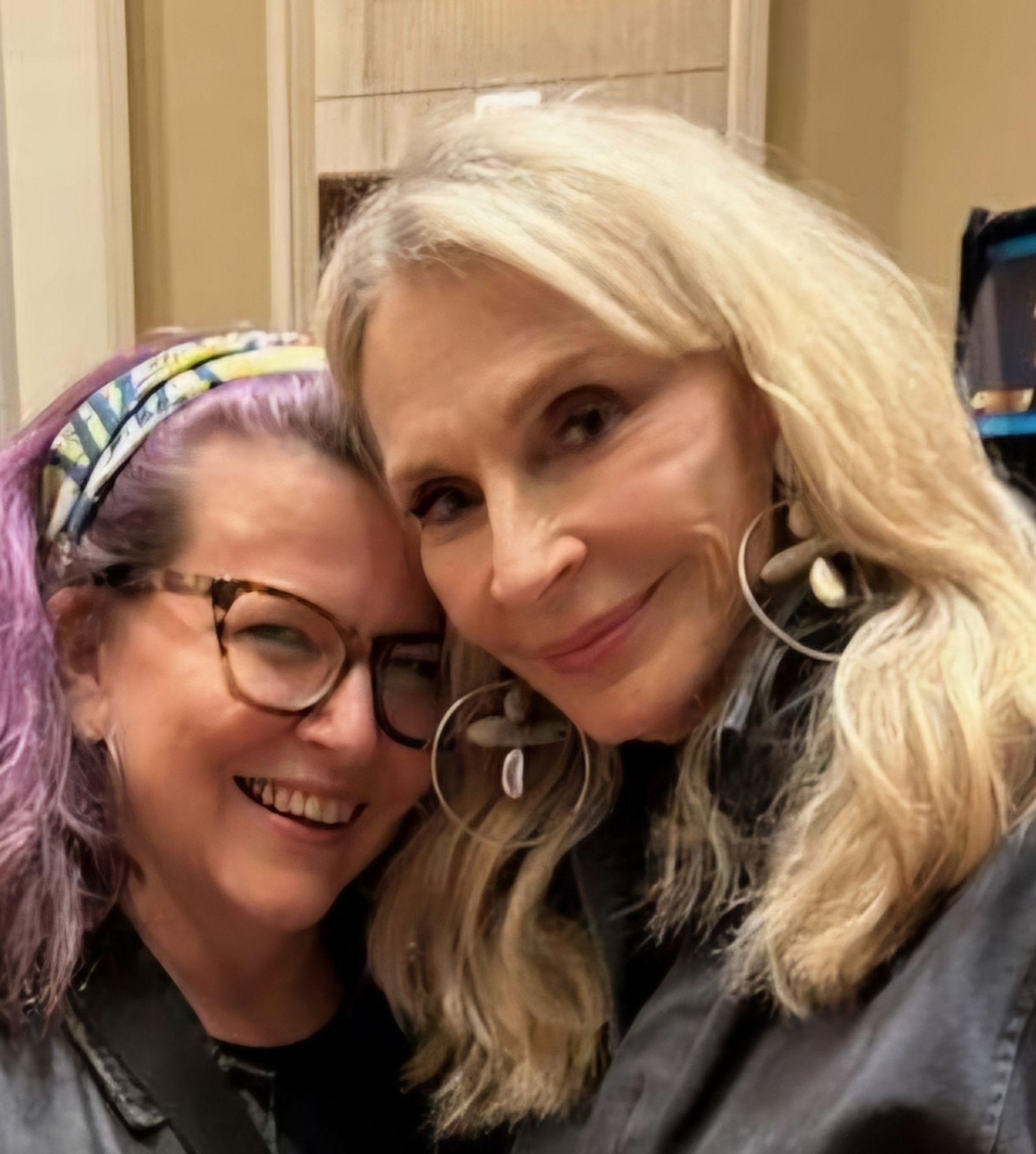 Two white women smiling with their heads close together; one has purple hair & tortoise shell glasses & is me; the other has long light blonde hair & big earrings & is Gates McFadden.