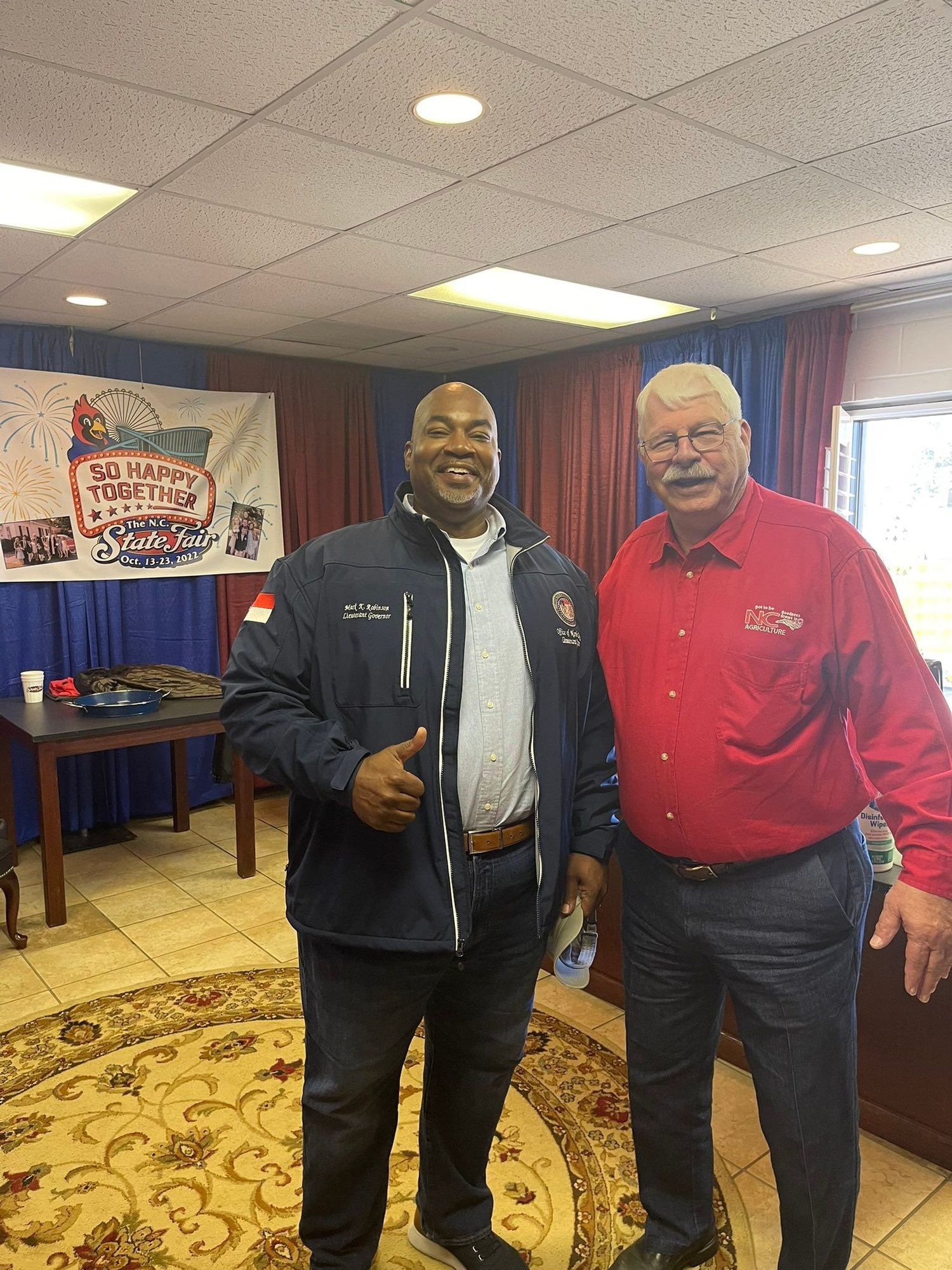 Mark Robinson and Steve Troxler standing together in front a banner that says "So Happy Together"