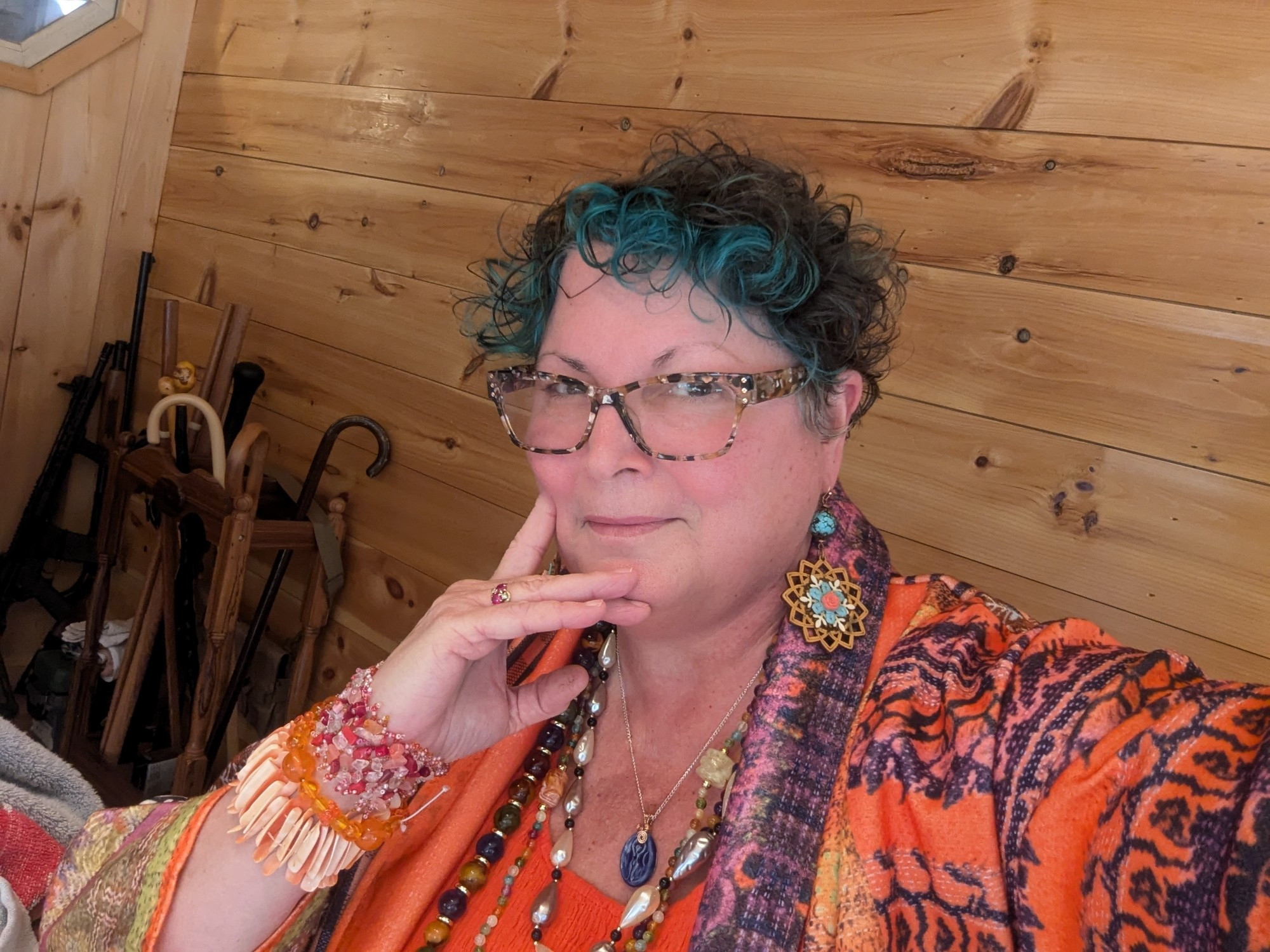 Photo of woman with blue hair wearing an orange dress, a multi-colored and patterned jacket with chunky orange and red bracelets and large beaded necklaces in purple, orange, and blue stones. Earrings are turquoise colored beads and wood.