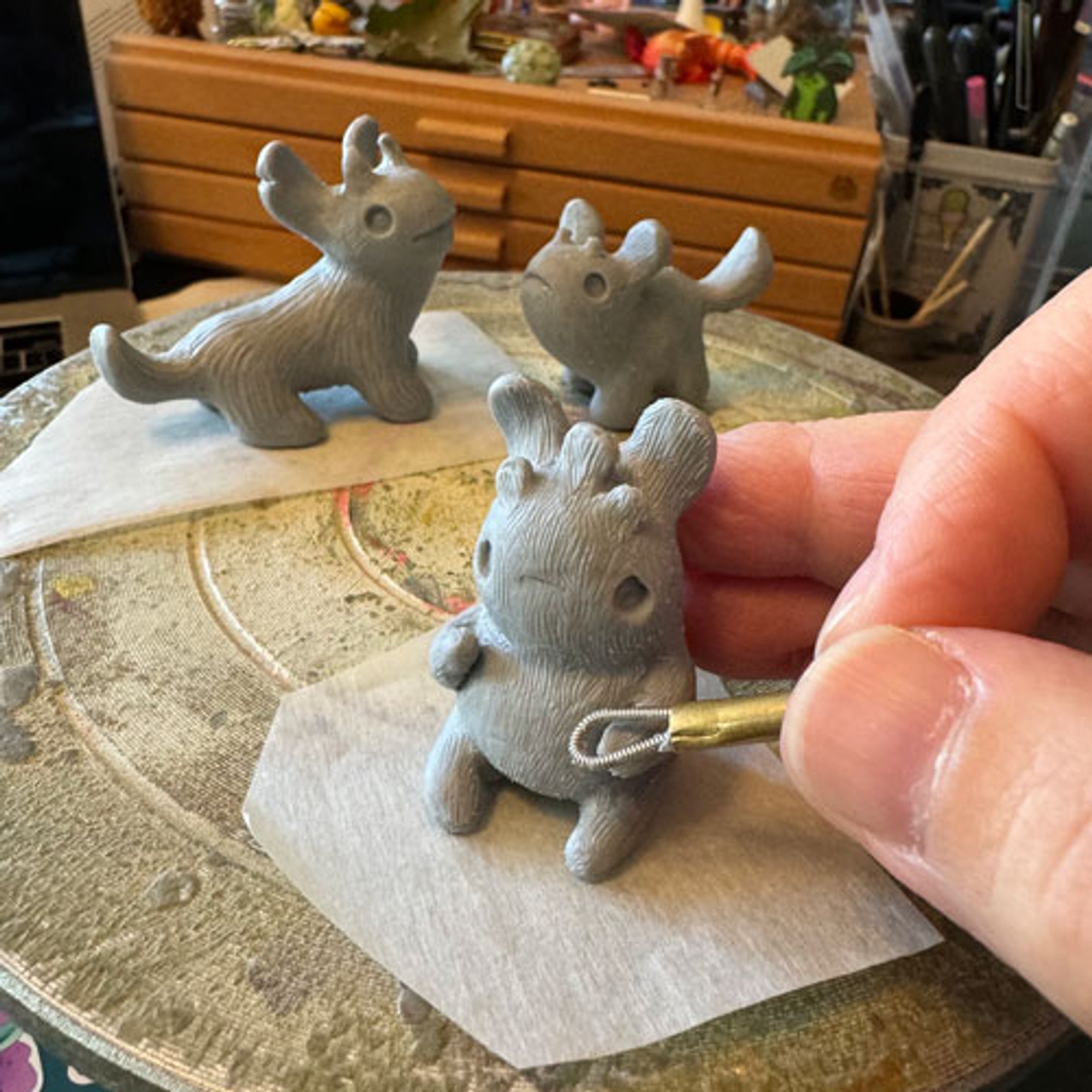 work in progress: grey polymer clay sculpts, with a hand brushing one with a wire tool. The sculpts are little creature dudes.