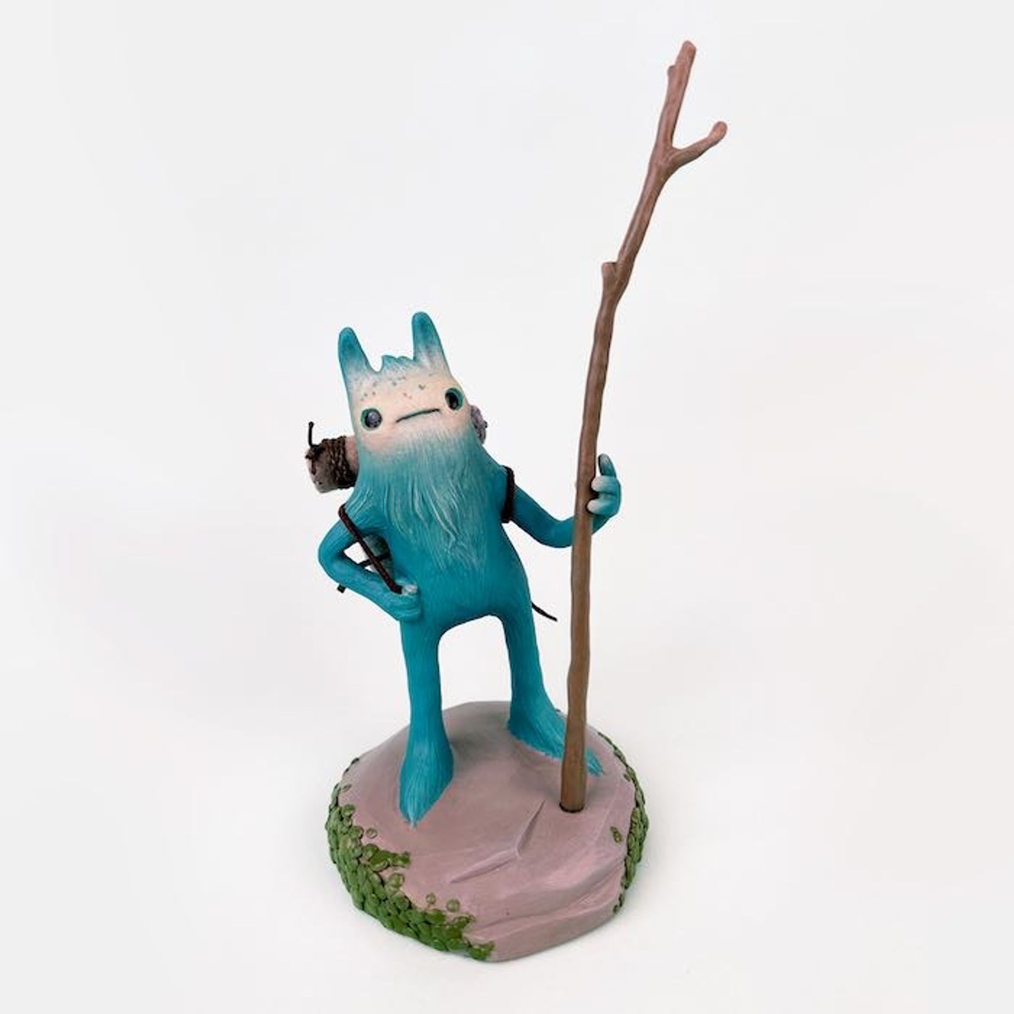 a jaunty little blue creature on a stone base (it's all sculpted) holding a twig (also sculpted because I'm an idiot) with a backpack and an adventurous nature.