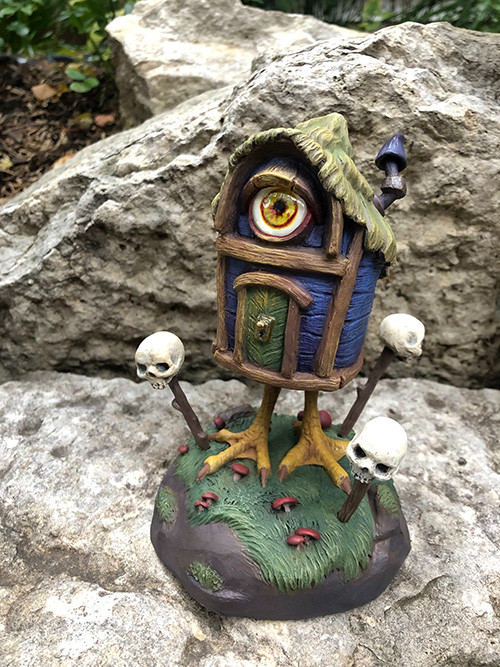 A sculpture sitting on craggy grey rocks. The sculpt is a blue-purple Baba Yaga hut with a thatched roof and three tiny skulls on sticks surrounding it. It stands on yellow chicken feet in a patch of grass with some red mushrooms growing around. The eye is a big glass bit over the door, it's yellow (watercolor!), and the door is green. Cute and menacing. 